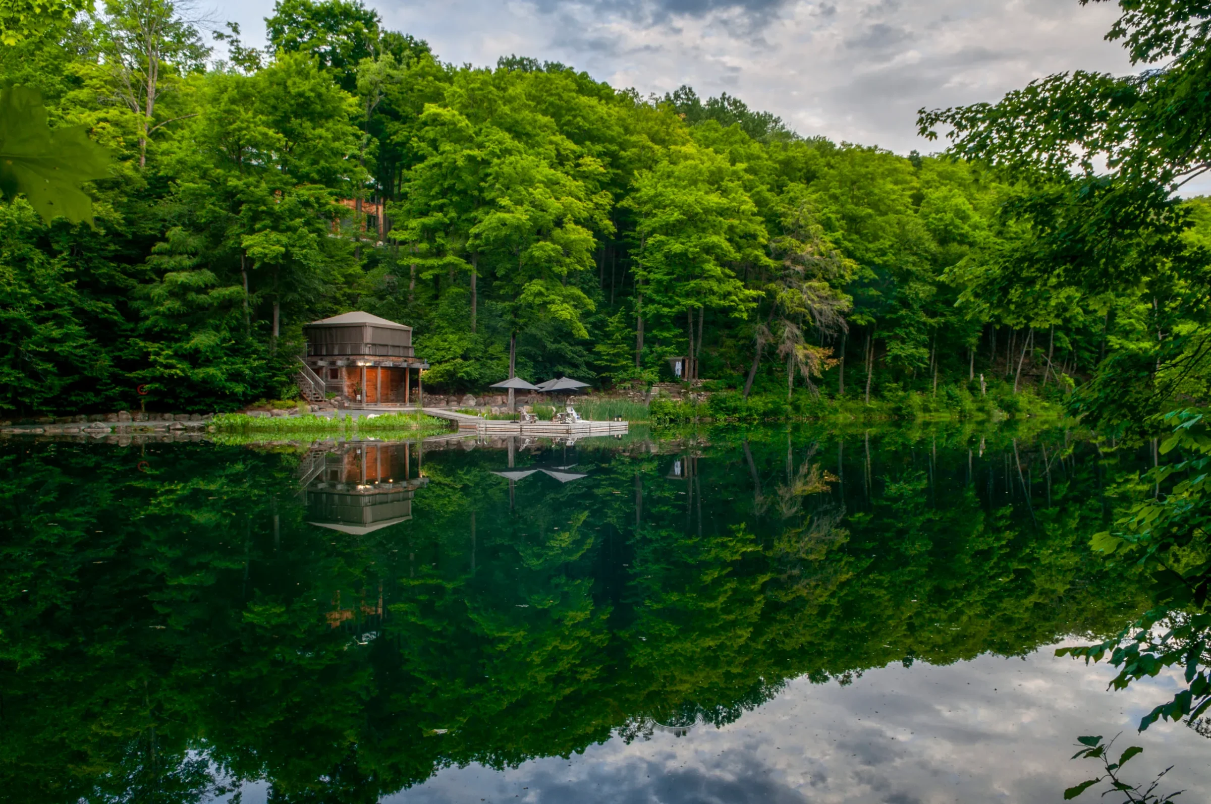 A serene forested lake scene features a wooden cabin on the shore, with reflections in calm water and lush green trees surrounding.