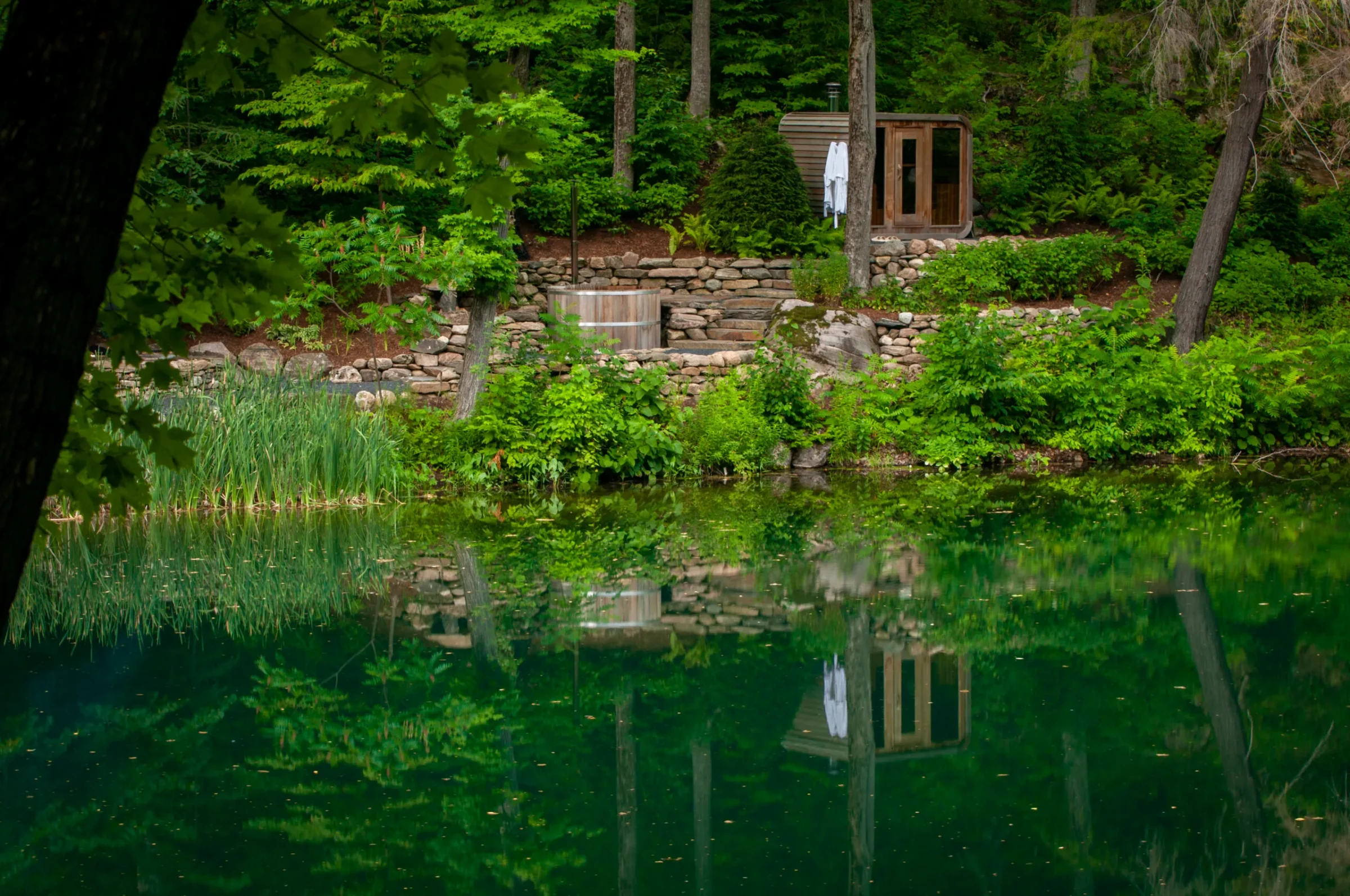 A serene pond reflects lush greenery, a small wooden structure, and a robe, creating a tranquil, natural atmosphere in a forested setting.