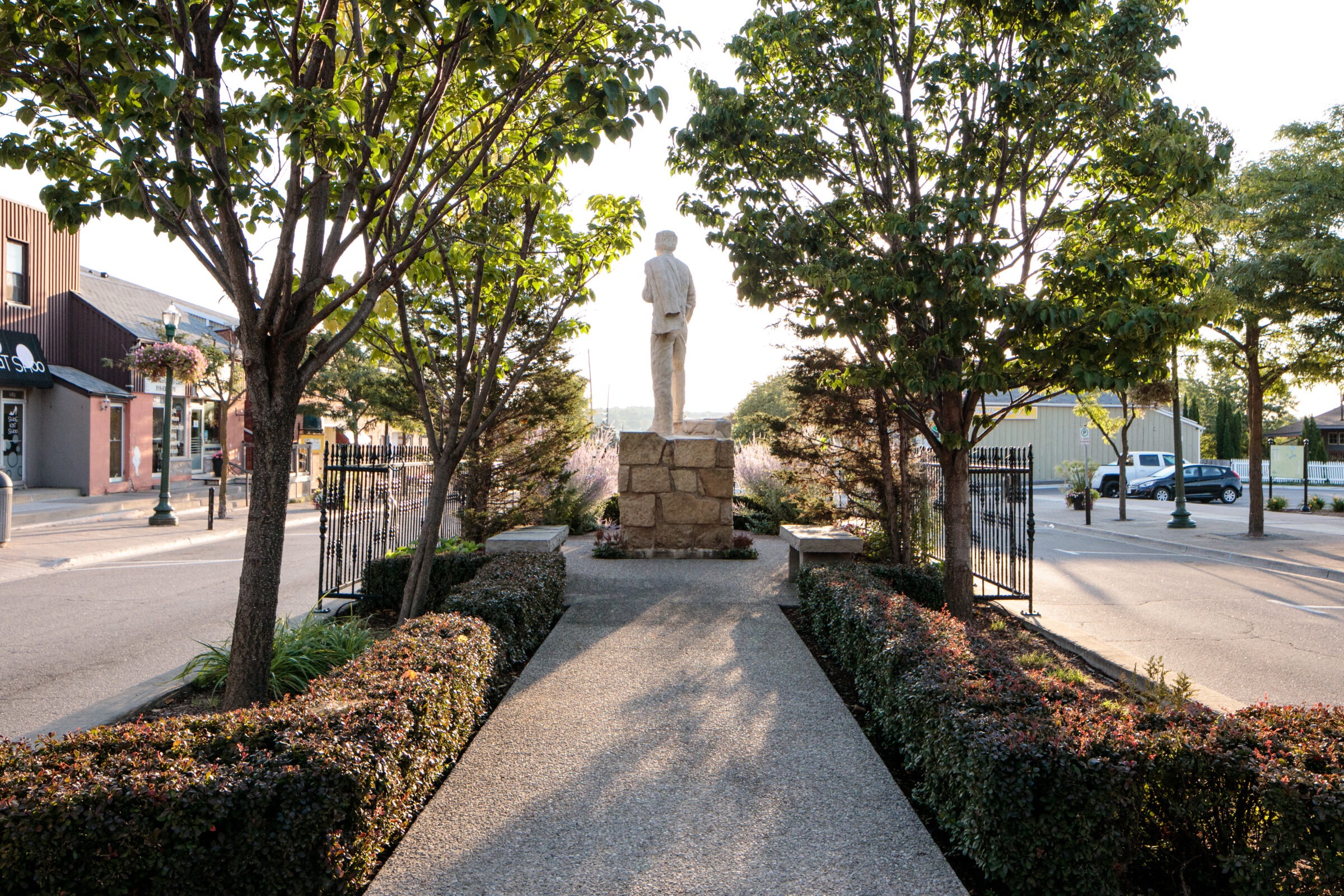A statue stands in a tree-lined walkway, with adjacent shops, benches, iron gates, and a street illuminated by setting sunlight.