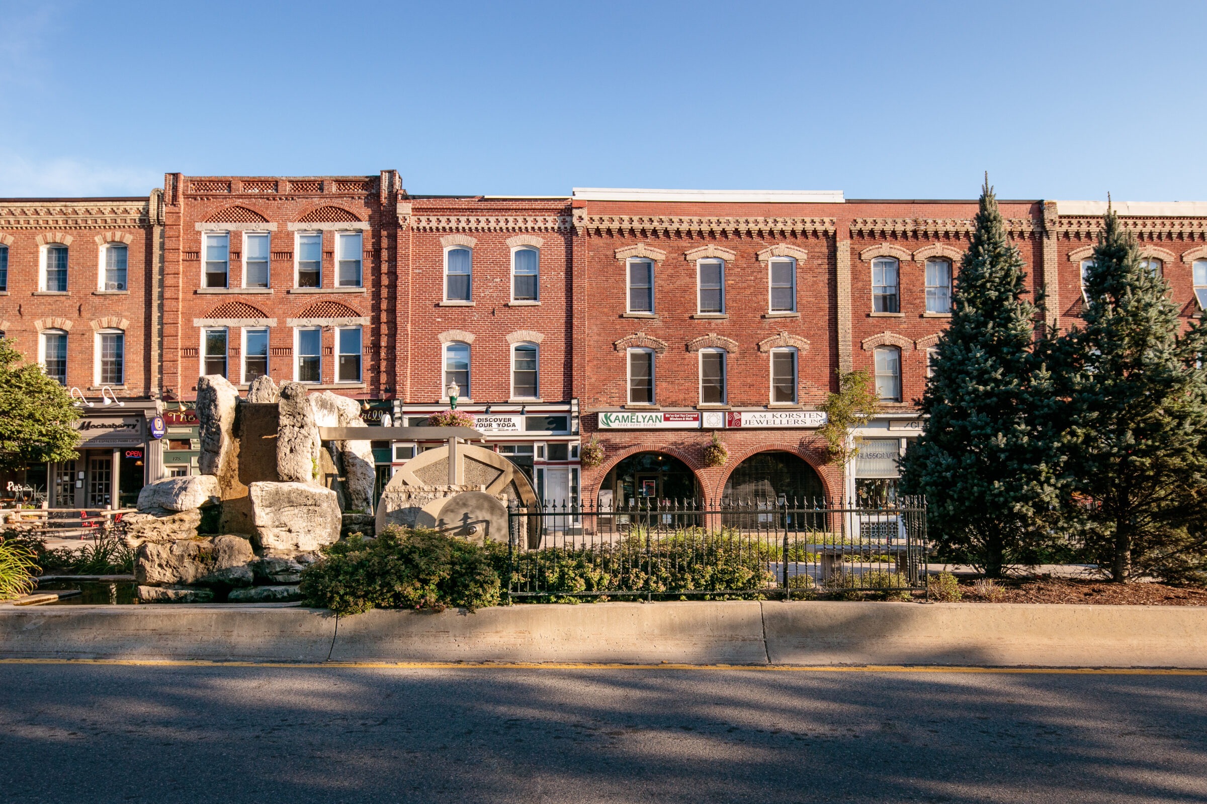 Historic red brick buildings with arched windows, storefronts, and decorative stone fountain. Evergreen trees and clear blue sky enhance the charming scene.