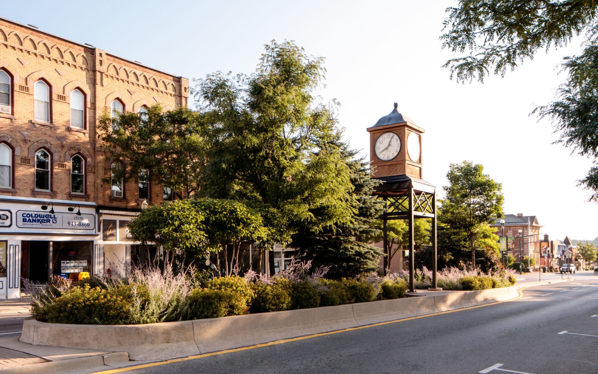 A street scene features a historic brick building, lush greenery, and a prominent clock tower under a clear blue sky.
