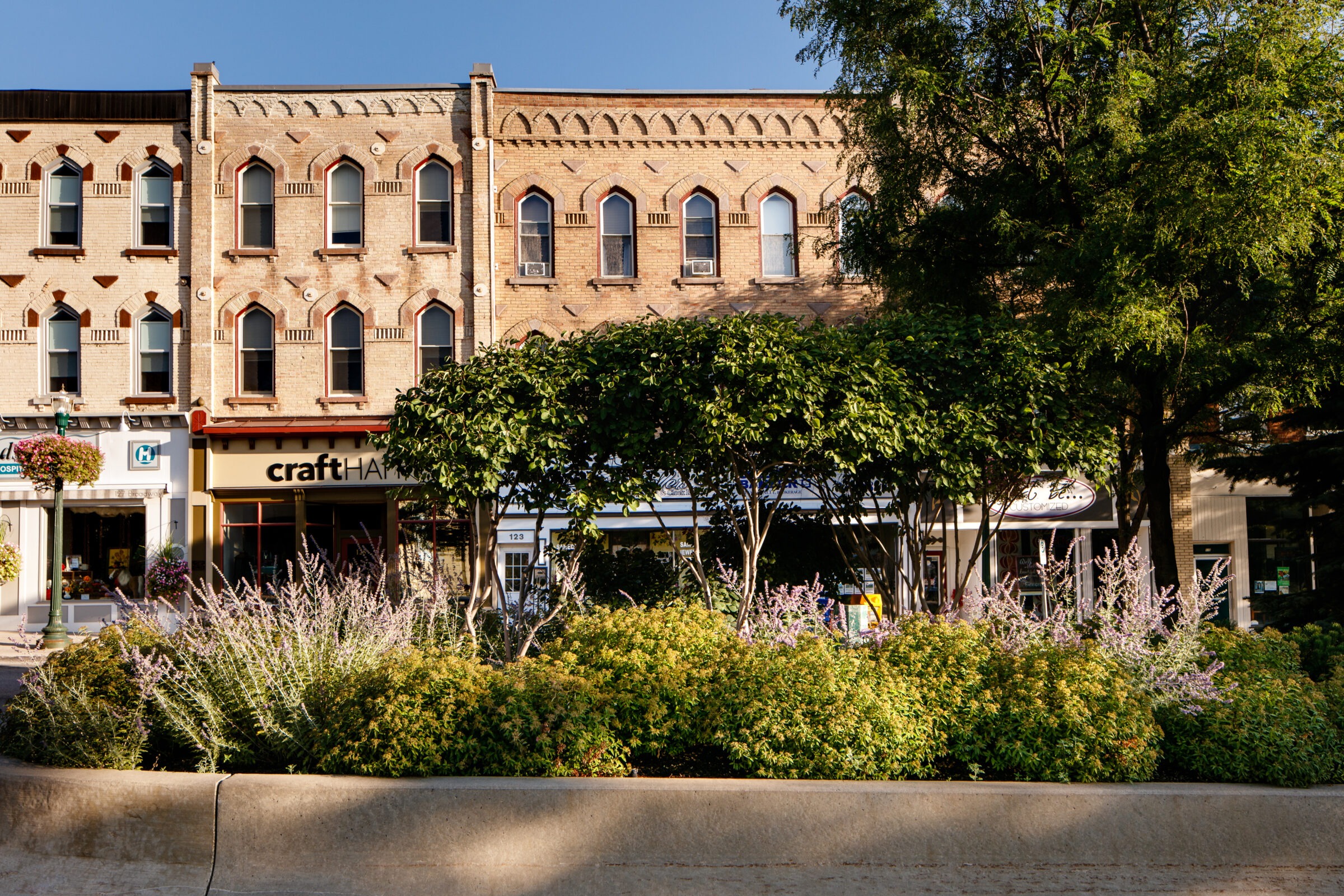 Historic building facade with arched windows, lined with lush trees and plants. Storefronts below include a craft shop, creating a charming street scene.