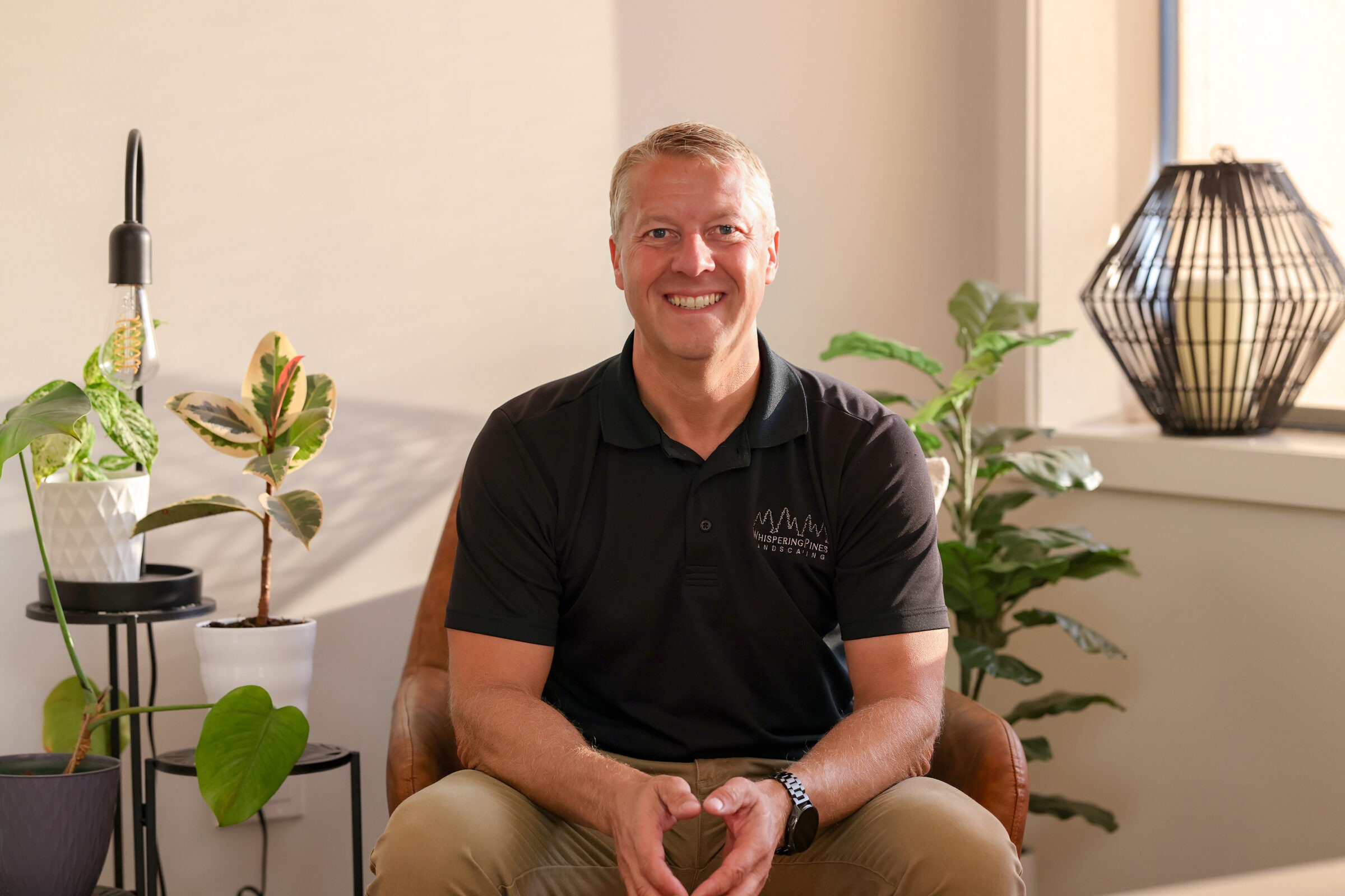 A person sits on a chair indoors, smiling, surrounded by potted plants and decorative lighting, in a well-lit, modern room.