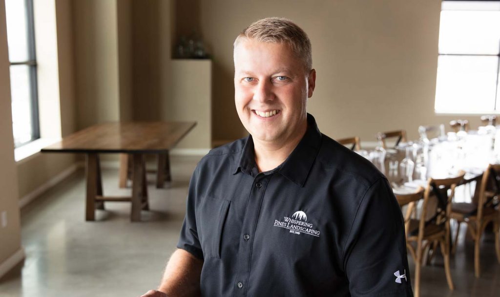 A person smiles in a well-lit room wearing a "Whispering Pines Landscaping" shirt, with a wooden table and chairs in the background.
