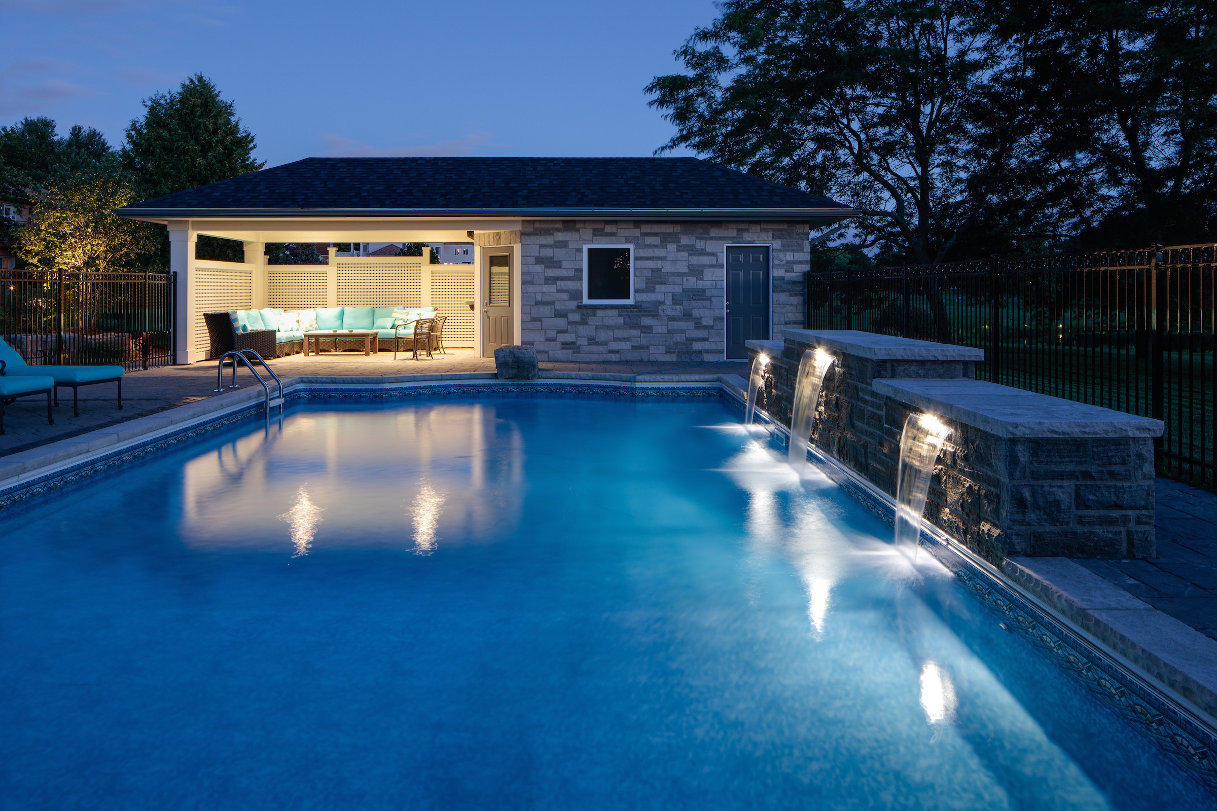 A serene backyard pool at dusk features cascading water, cozy seating under a gazebo, and softly lit surroundings, creating a tranquil atmosphere.