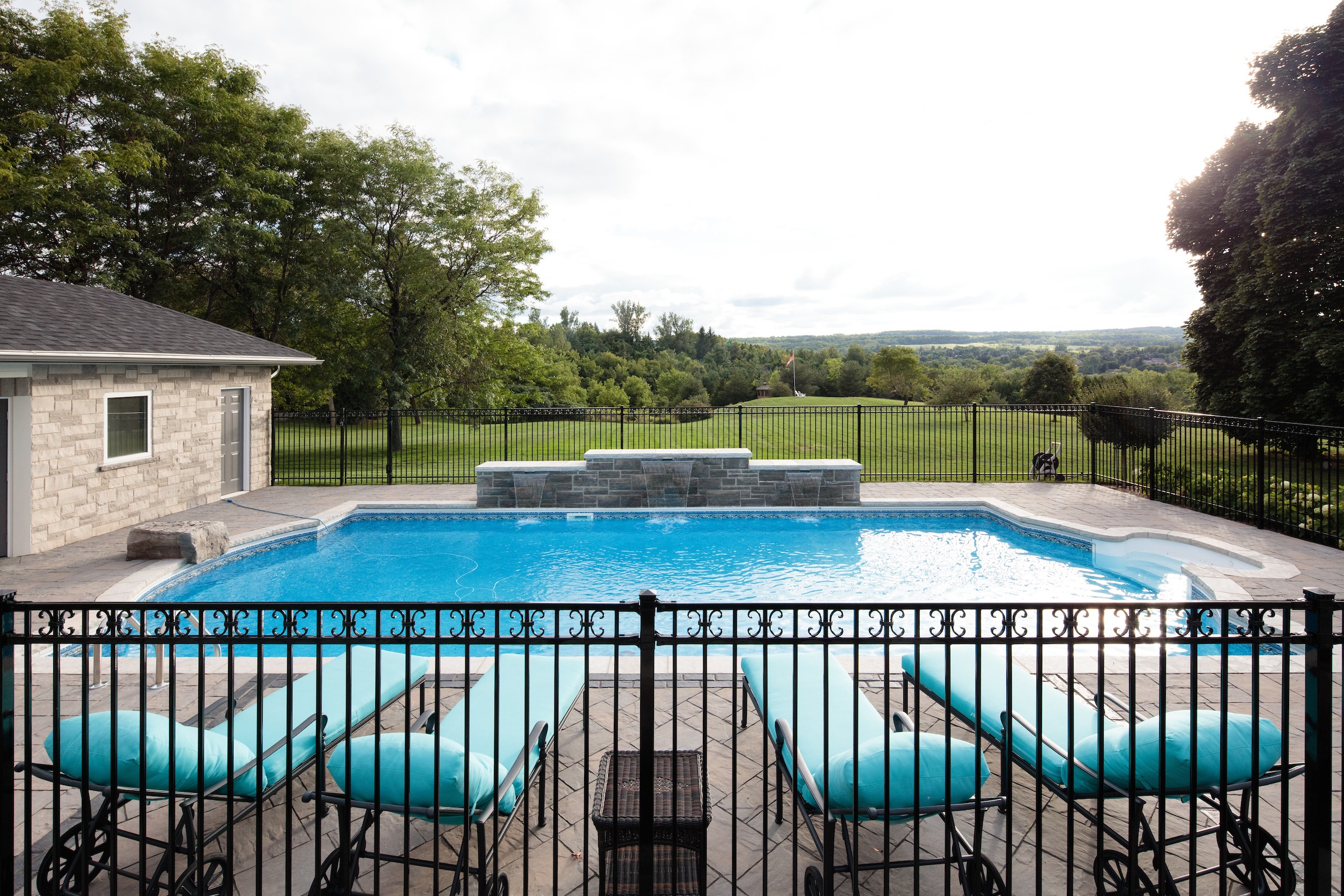 A serene backyard features a pristine swimming pool, loungers, and lush greenery, overlooking a scenic landscape under a partly cloudy sky.