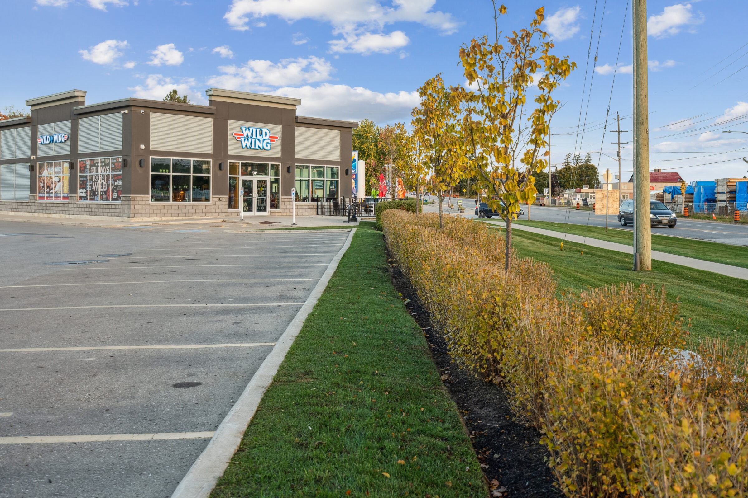A "Wild Wing" restaurant is beside an empty parking lot. Trees and bushes line the sidewalk under a partly cloudy sky.