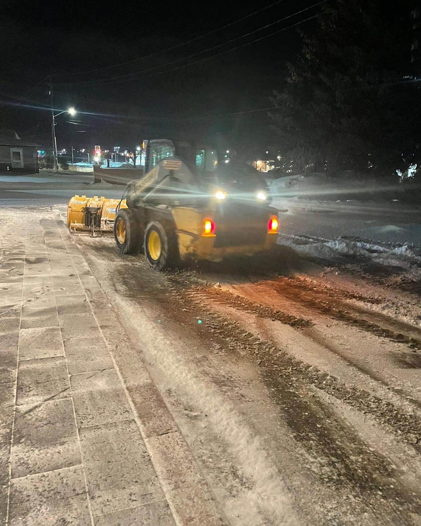 A snowplow is clearing snow-covered pavement at night, with bright streetlights illuminating the area. No recognizable landmarks or historical buildings are visible.