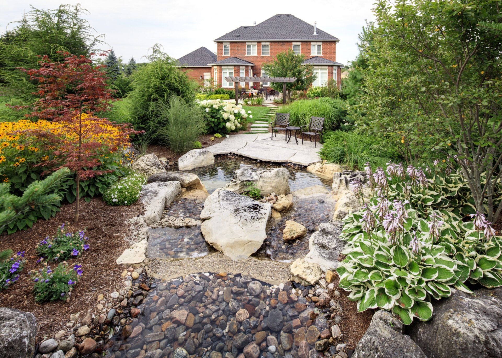 Lush garden with vibrant flowers and a small stream in the foreground, leading to a brick house with patio seating.