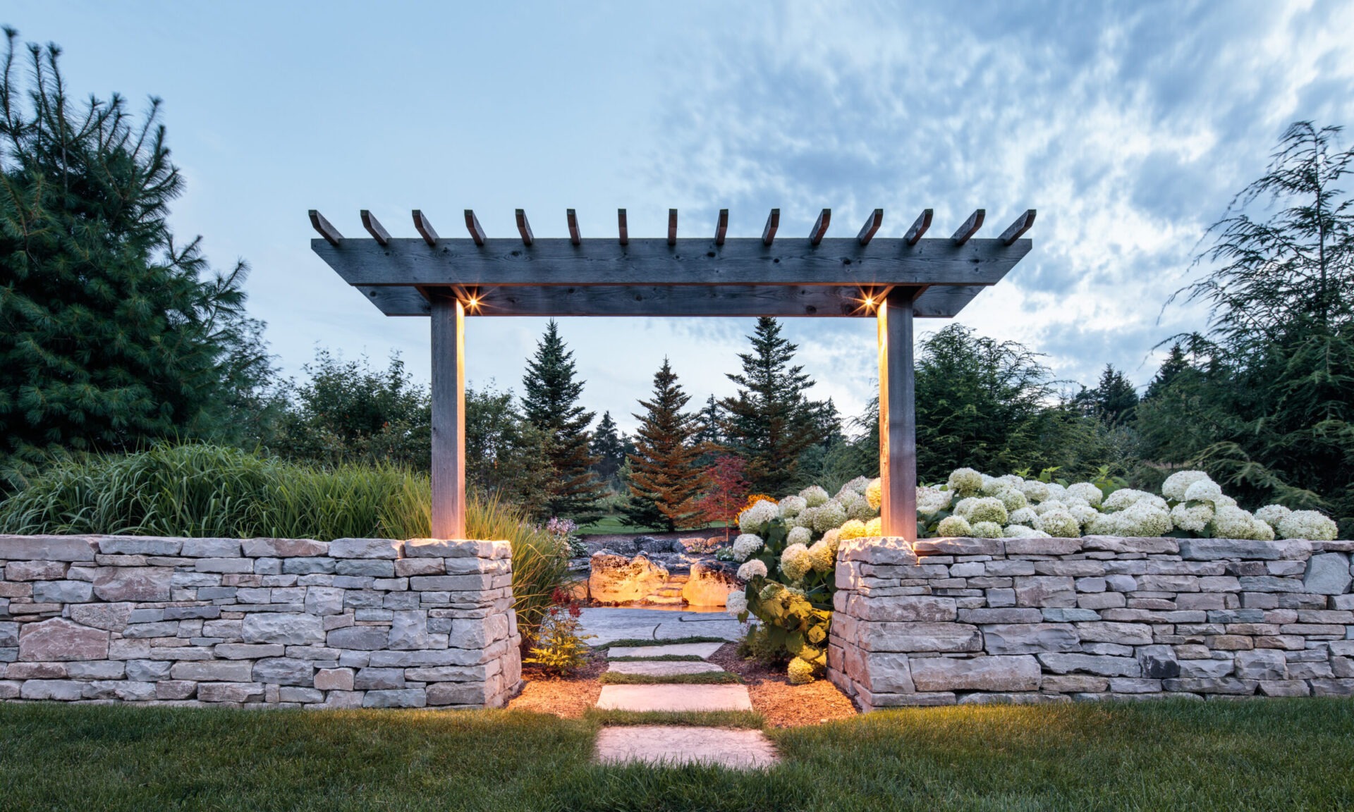 A stone pathway leads through a pergola surrounded by lush greenery and hydrangeas, with trees and a cloudy sky in the background.