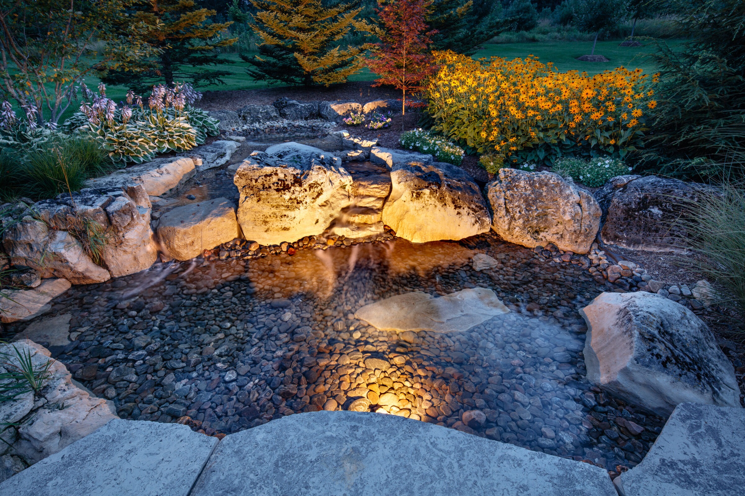 A small illuminated garden pond with rocks, surrounded by vibrant flowers and trees, creating a serene outdoor scenery at twilight.
