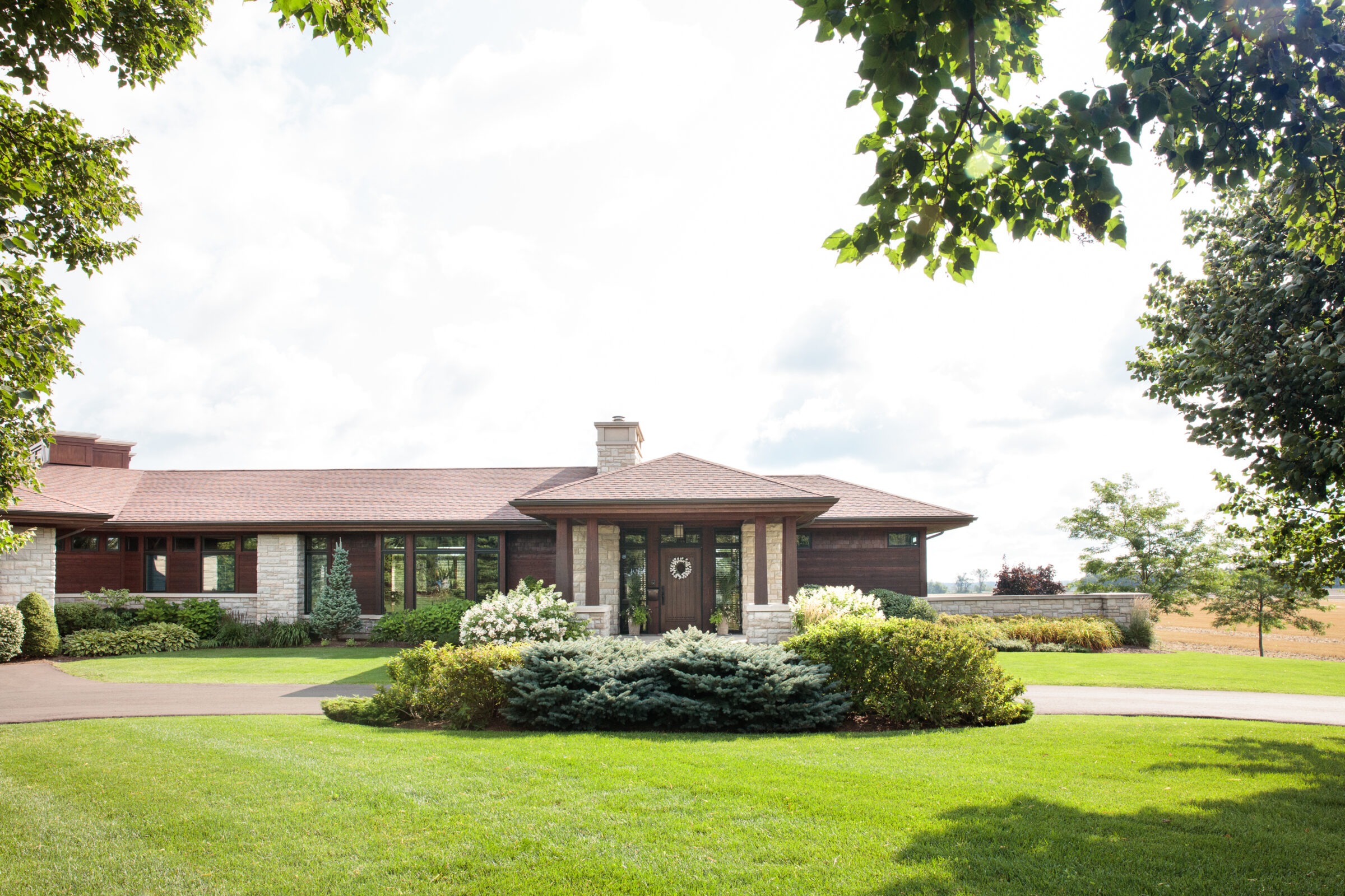 A modern, single-story house with a landscaped front yard, surrounded by trees and an expansive lawn under a partly cloudy sky.
