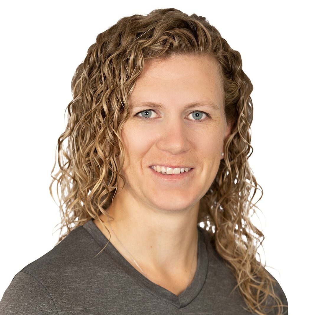 Person with curly hair smiles while wearing a gray "Whispering Pines Landscaping" shirt against a plain white background.