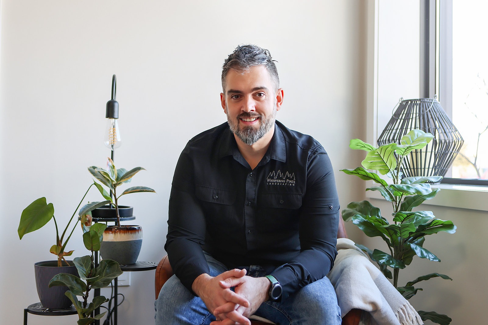 A person sits in a modern, plant-filled room with a light fixture. The person wears a "Whispering Pines Landscaping" shirt.