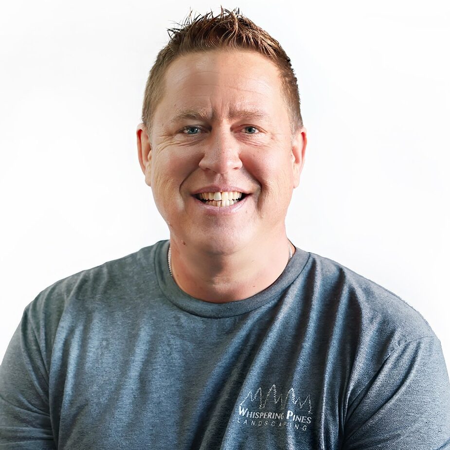 A person with short hair smiles, wearing a grey shirt reading "Whispering Pines Landscaping," against a plain white background. No landmarks visible.