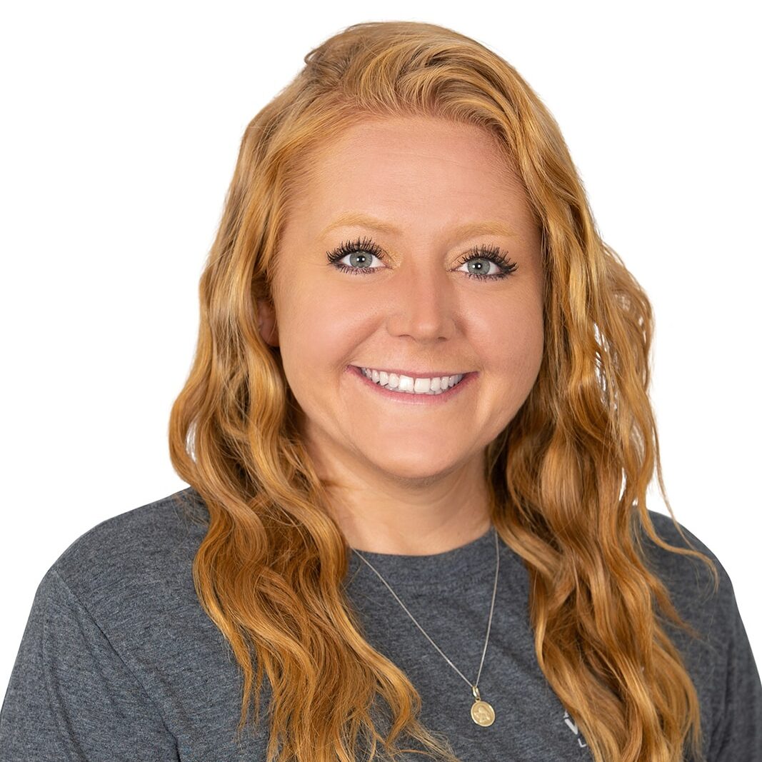 A person with long, wavy blonde hair and a friendly smile wears a gray t-shirt and a necklace against a plain white background.
