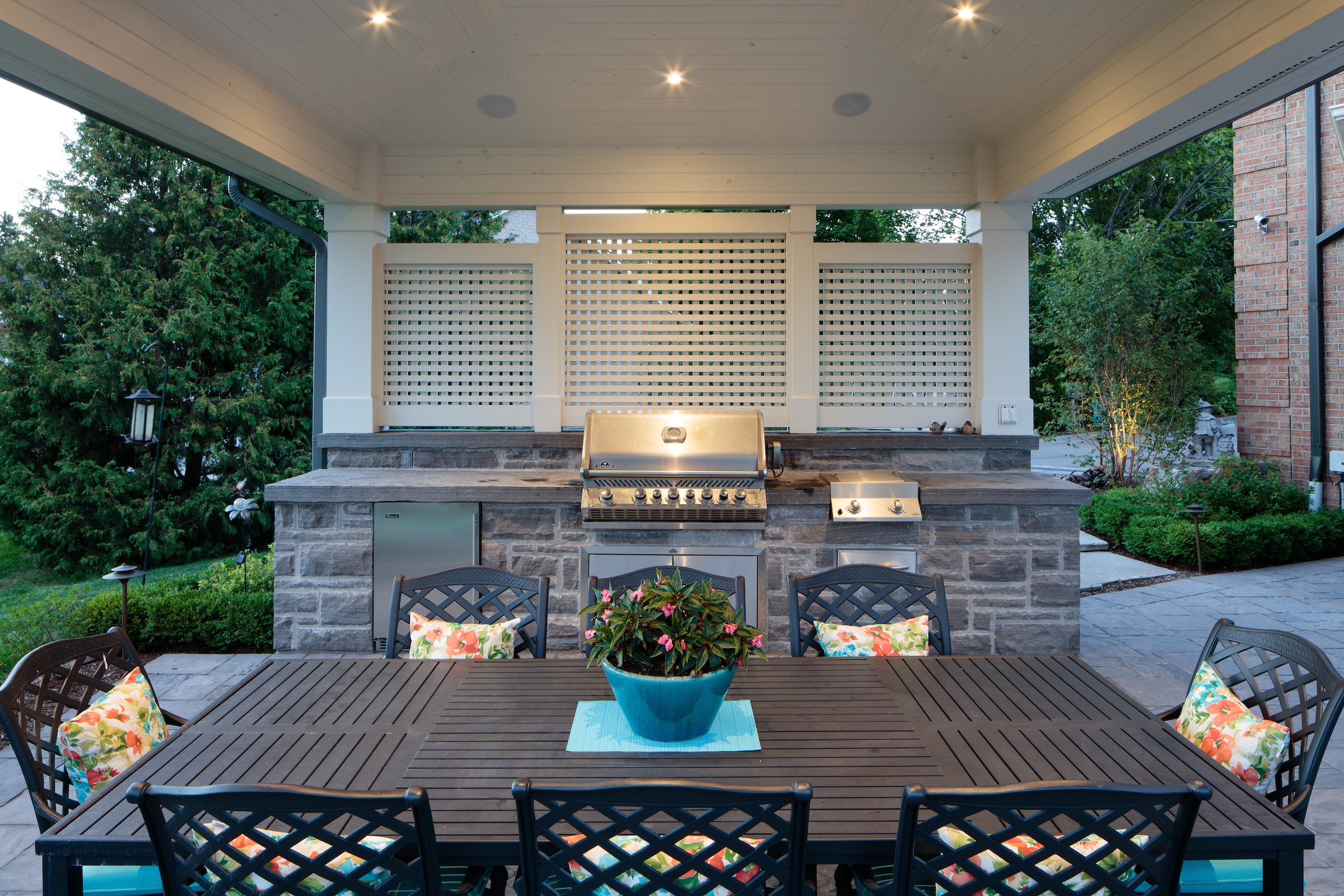 Outdoor patio with modern grill station, surrounded by greenery. Wooden dining table adorned with a potted plant and colorful cushions.