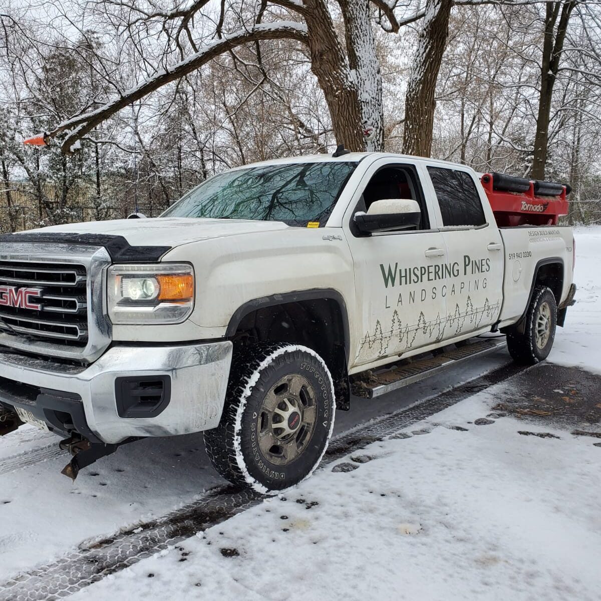 A white GMC truck with "Whispering Pines Landscaping" branding is parked on a snowy road, surrounded by snow-covered trees.