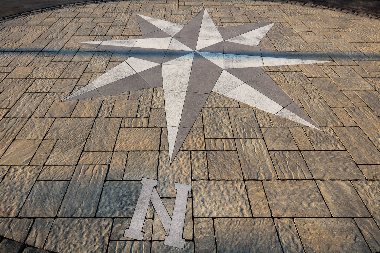A large compass rose embedded in a stone pavement with letter "N" for north, surrounded by textured rectangular tiles.