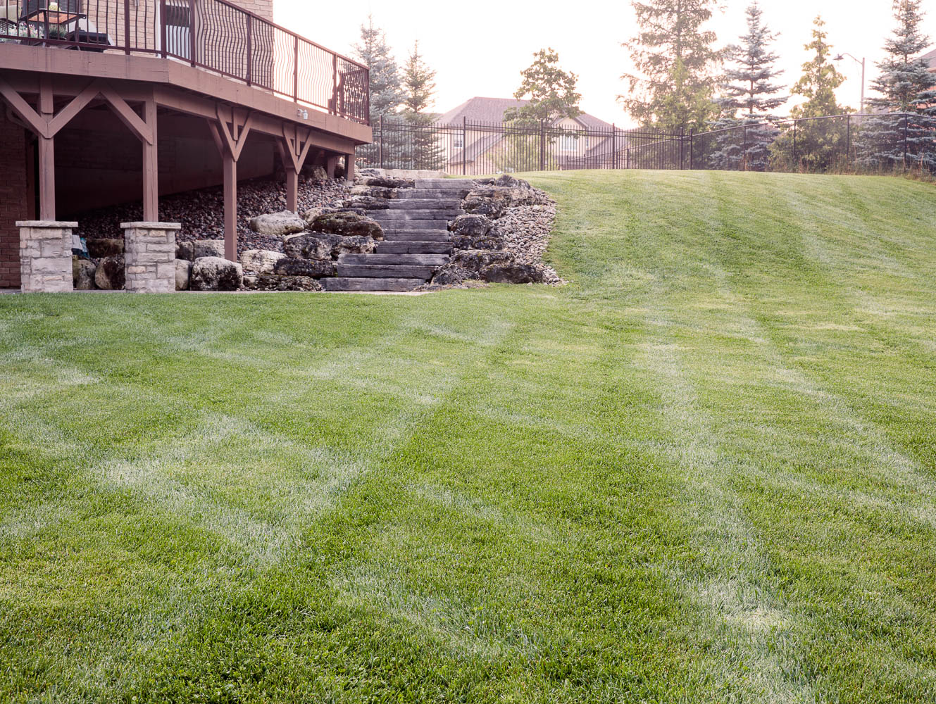 A well-maintained lawn with stone steps leads to a raised wooden deck, surrounded by trees and a fence in the background.