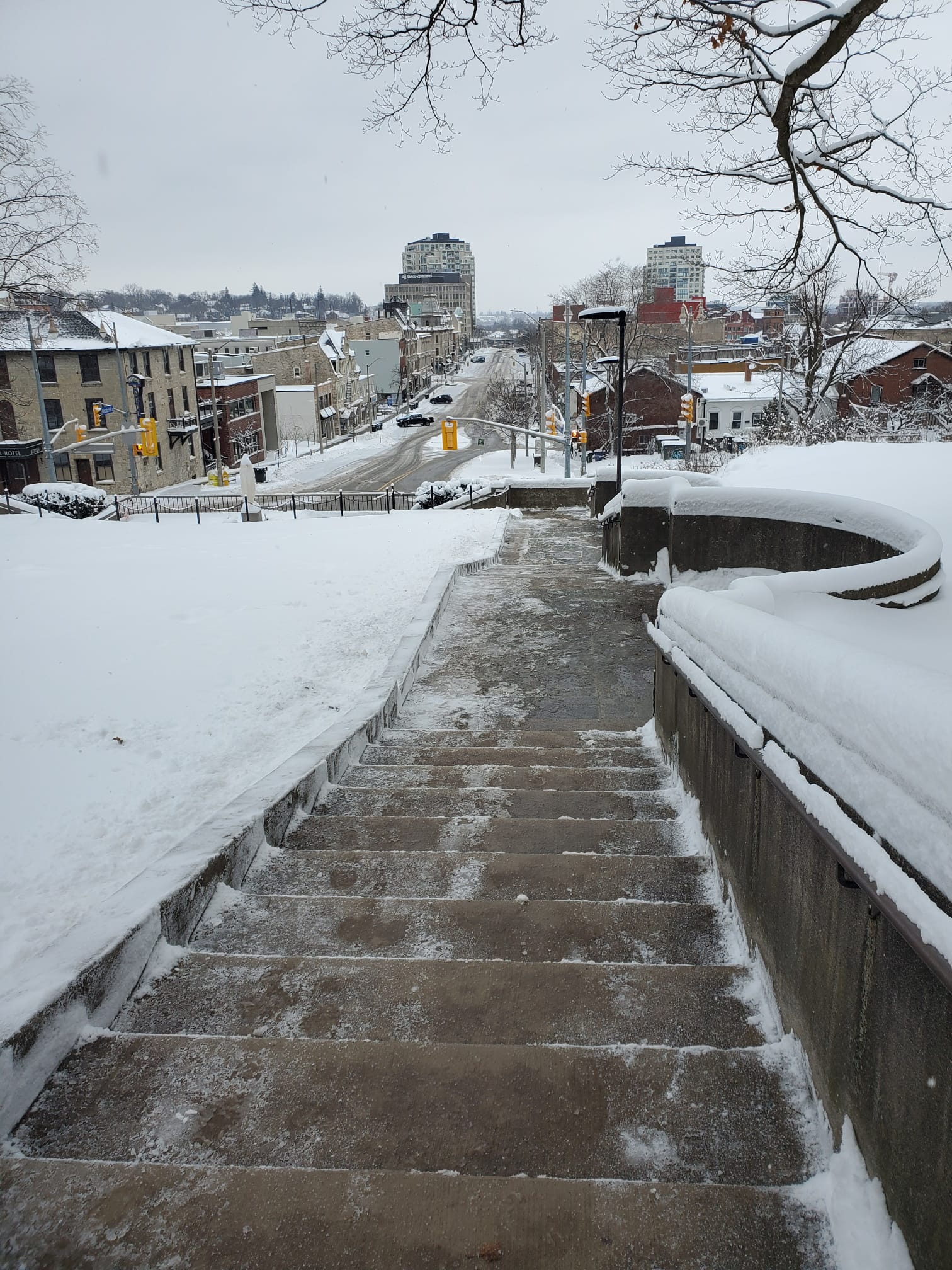 Snowy stairway leading to a street lined with buildings in a winter scene. Two tall buildings can be seen in the distance.
