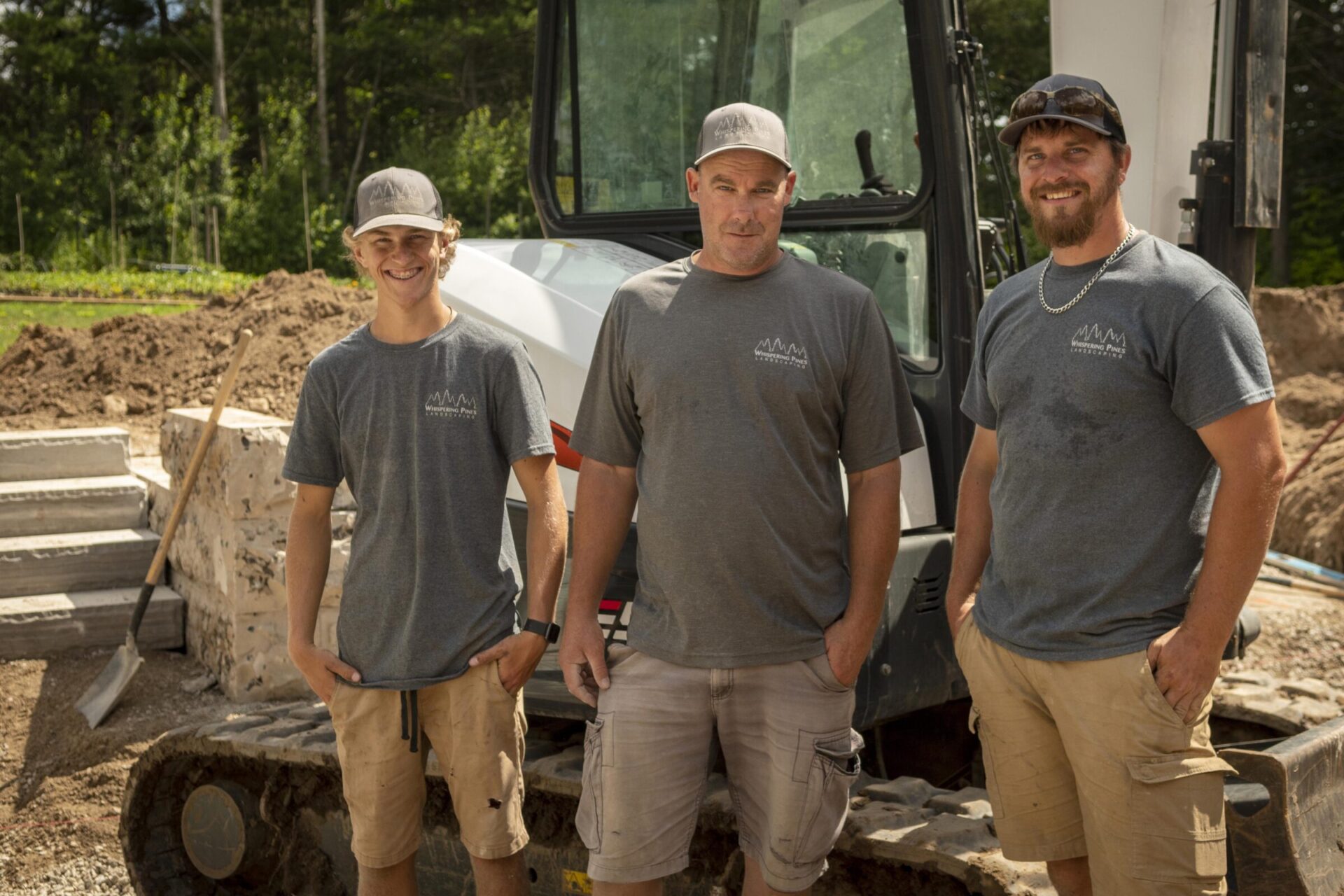 Three people wearing matching shirts and caps stand in front of a construction vehicle, surrounded by dirt and tools, outdoors.