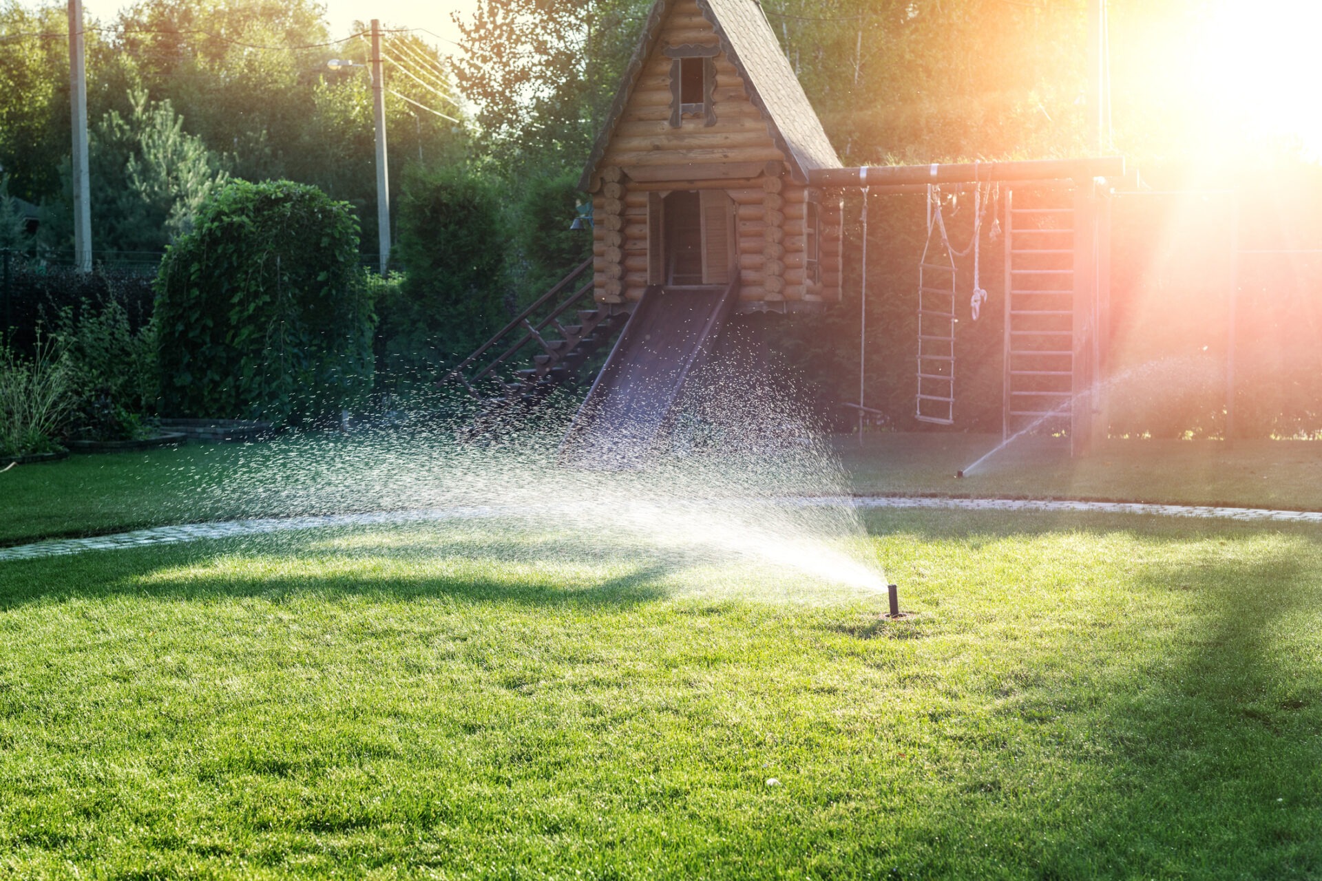 A sunlit backyard with lush grass, a wooden playhouse, and a sprinkler spraying water, surrounded by trees and climbing equipment.