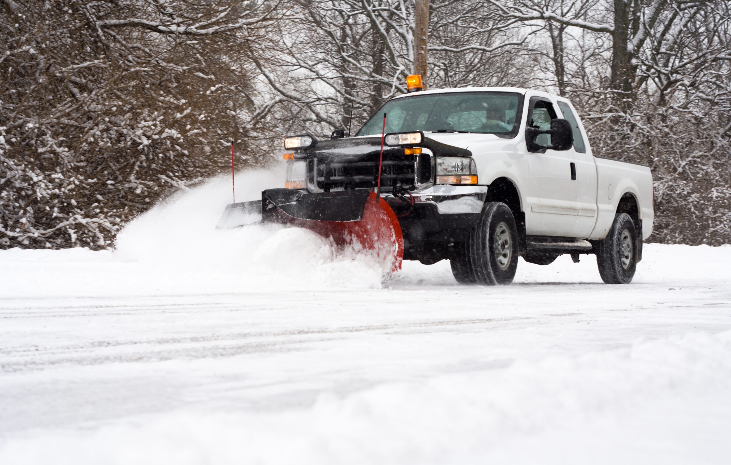 A snowplow truck clears snow on a wintery road surrounded by leafless trees, under a gray sky, creating piles of snow on the sides.