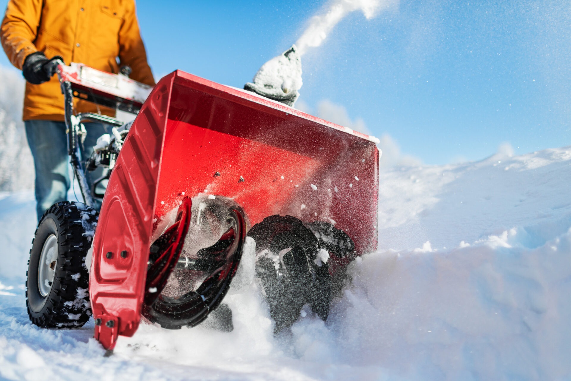 A person operates a red snowblower, clearing thick snow under a bright blue sky, with snow flying through the air.