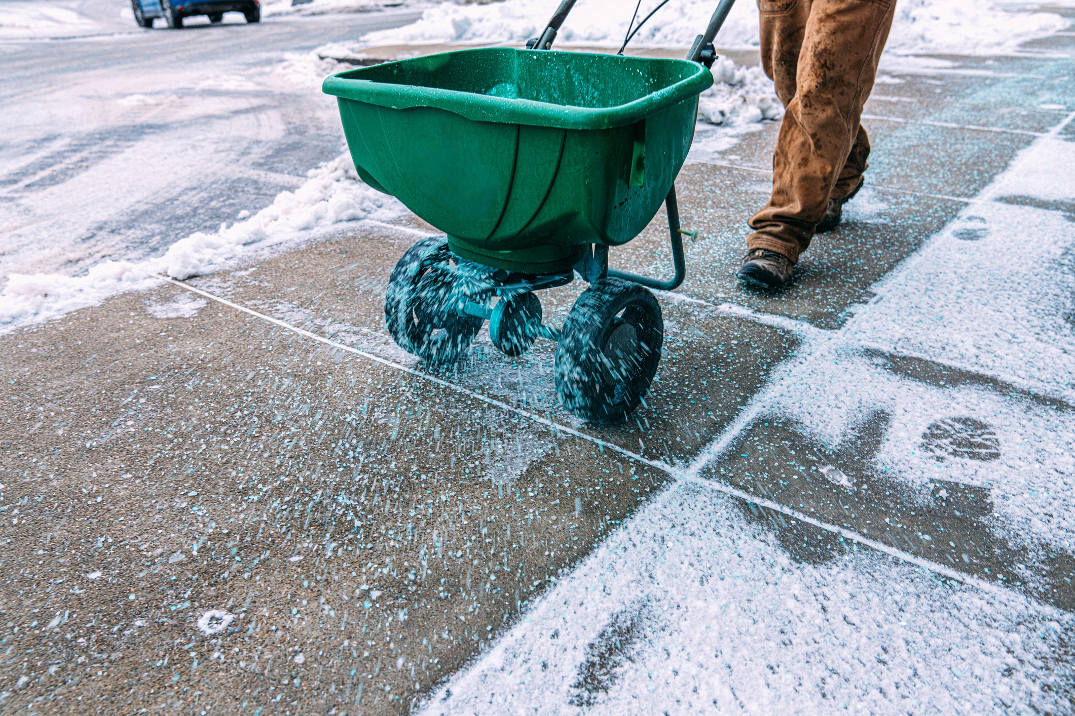 A person pushes a green spreader, dispersing salt on a snowy sidewalk to prevent ice accumulation, enhancing pedestrian safety in winter.