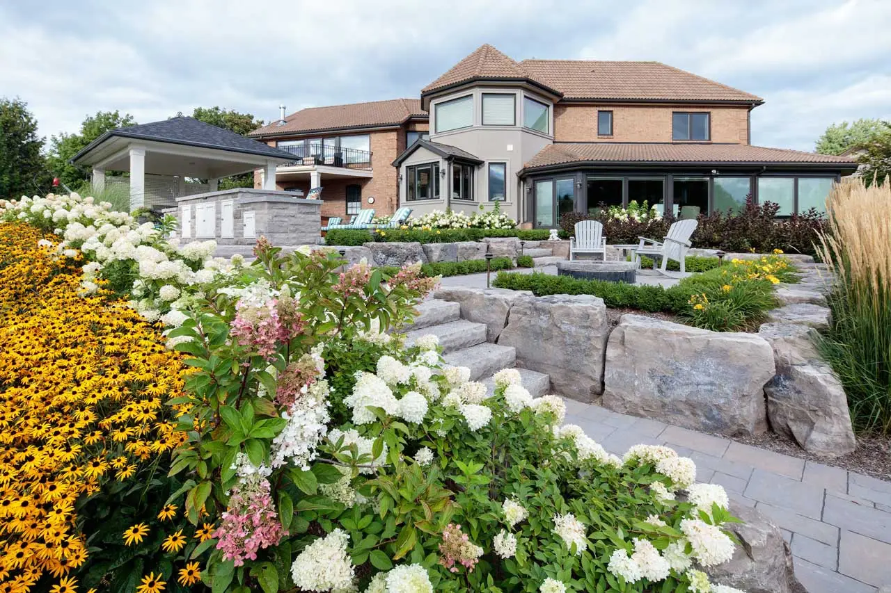 A large modern house with a landscaped garden, featuring colorful flowers, stone steps, and outdoor seating under a cloudy sky.