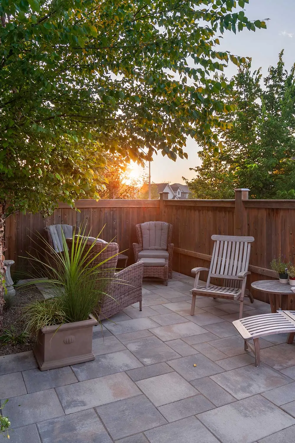 Cozy patio with wooden chairs and potted plants, surrounded by a wooden fence, under leafy trees. Sunsets in the background.