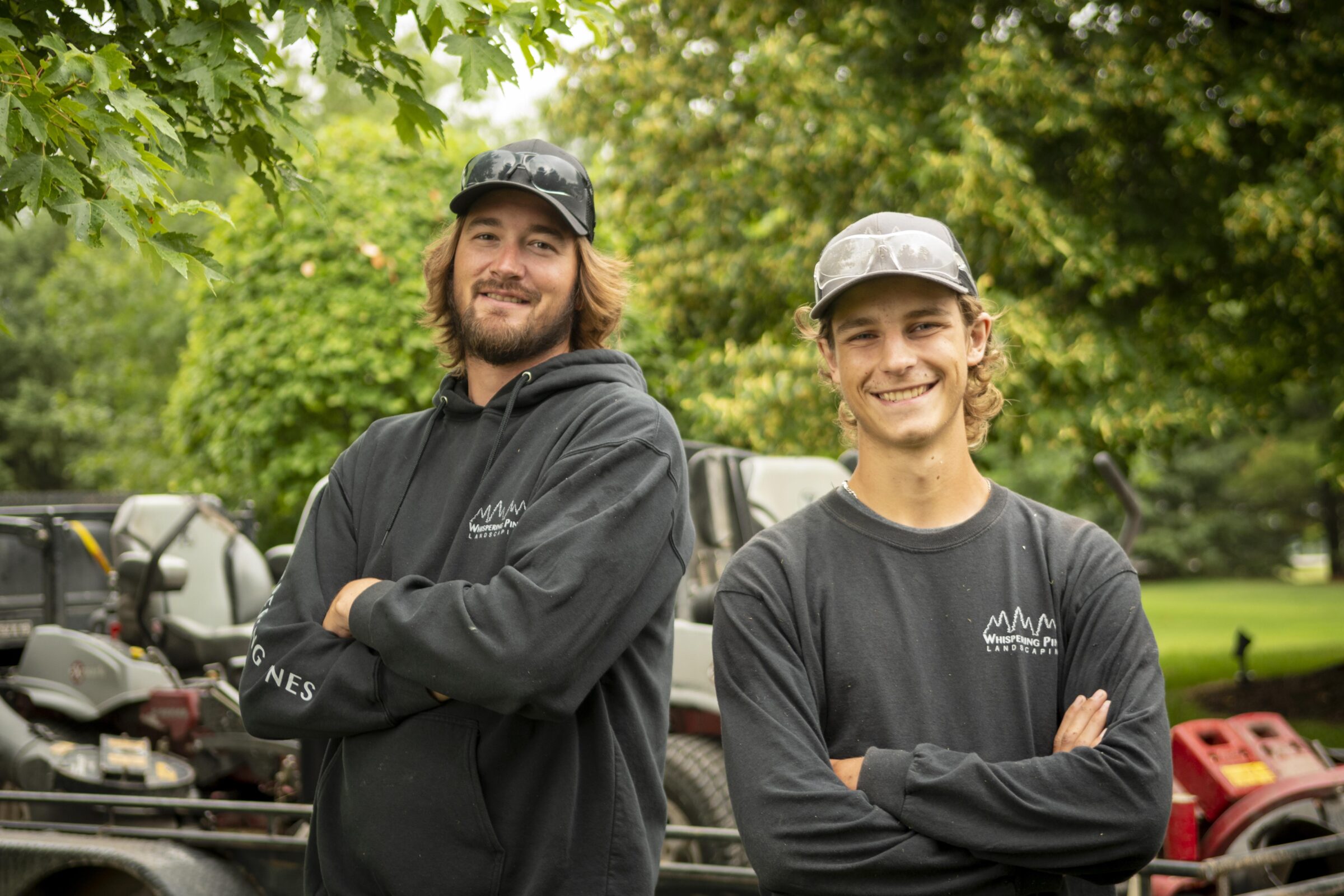 Two people standing outdoors with arms crossed, near garden equipment and lush green trees, wearing casual work attire and caps.