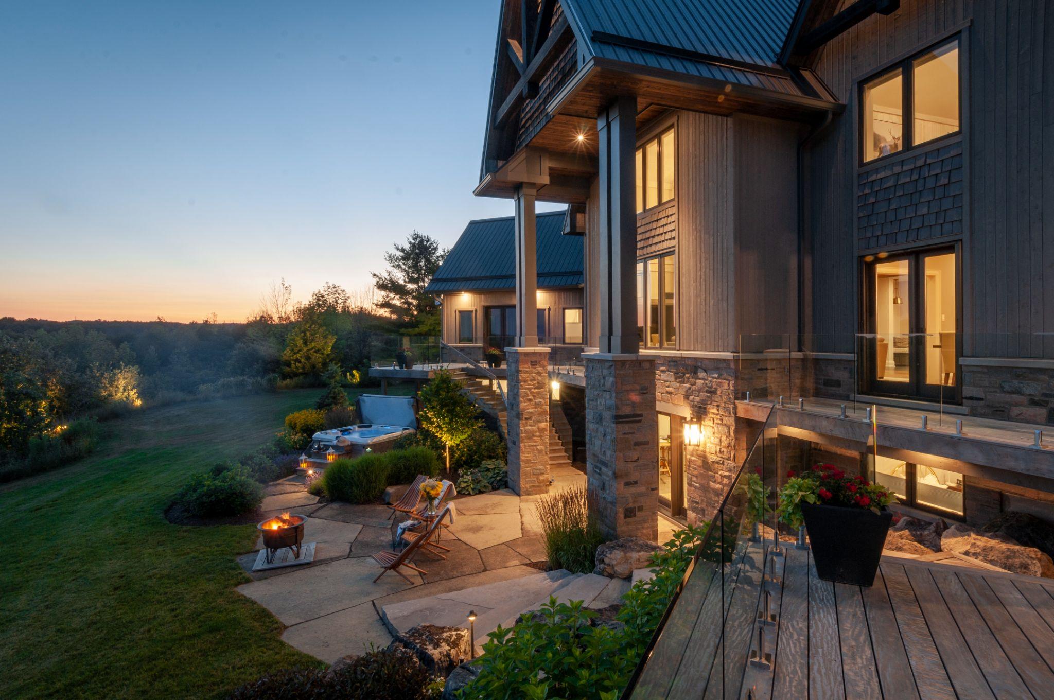 An inviting evening view of a modern home illuminated by warm lights, highlighting the outdoor living space with a fire pit and lounge chairs, set against a backdrop of lush greenery and a gradient sunset sky.