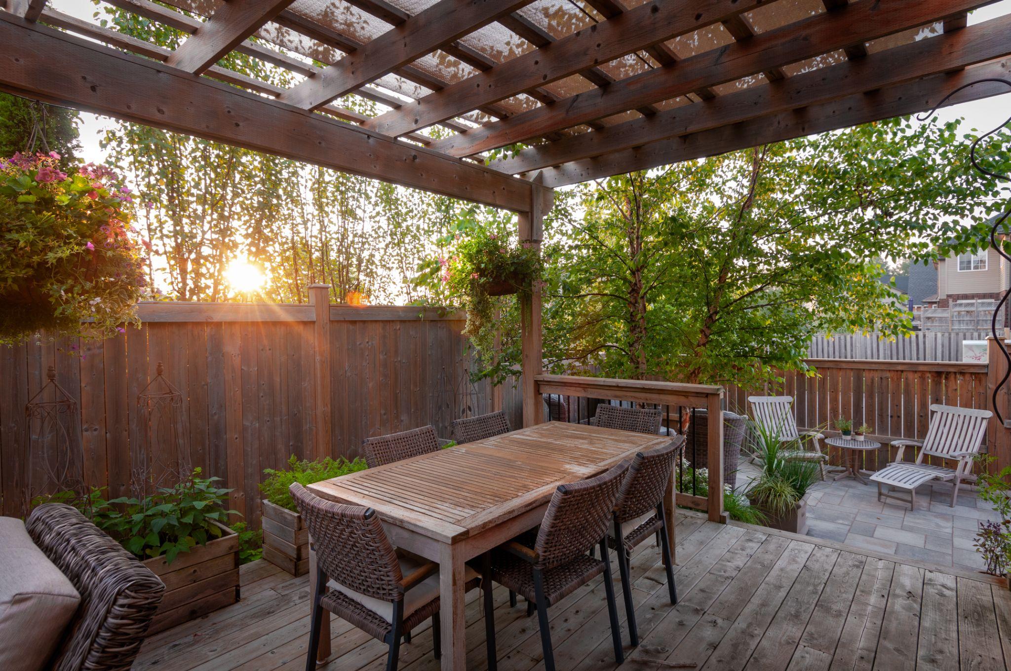 A charming outdoor dining area under a wooden pergola, featuring a large table surrounded by wicker chairs, with hanging plants and a beautiful sunset illuminating the space, set against a backdrop of greenery and wooden fencing.