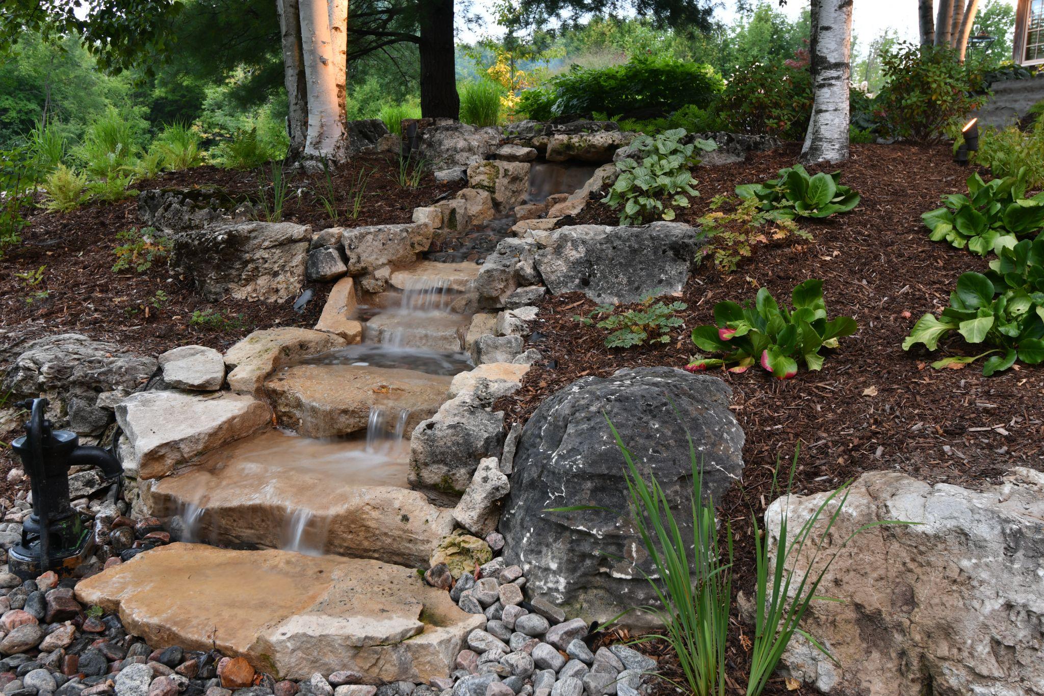 A beautiful garden scene featuring a cascading water feature flowing over natural stone steps, surrounded by lush green plants and trees, with a rustic pump in the foreground.
