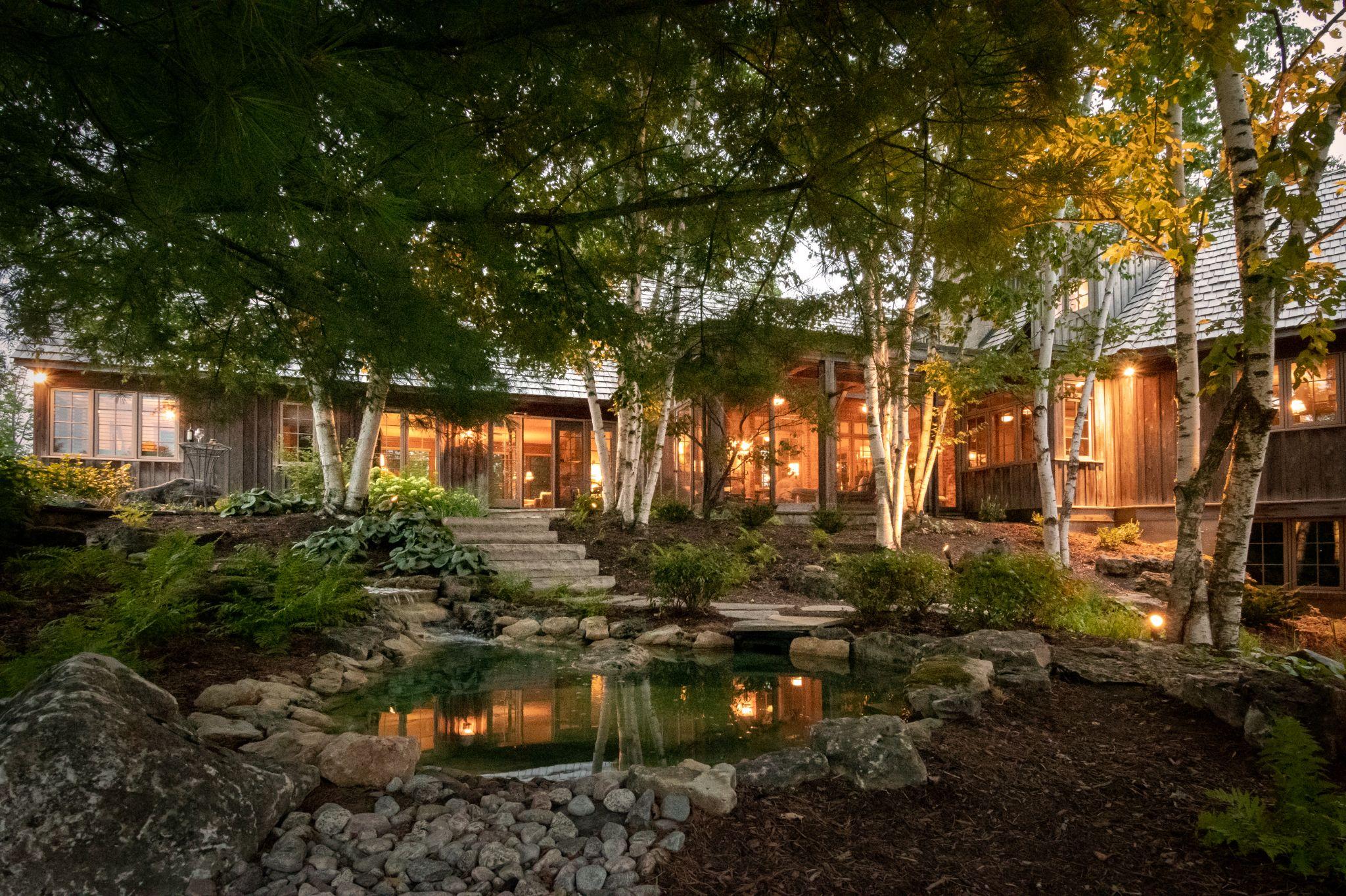 A cozy wooden house surrounded by lush greenery and illuminated by warm lights, featuring a tranquil pond with smooth stones in the foreground and birch trees framing the scene.