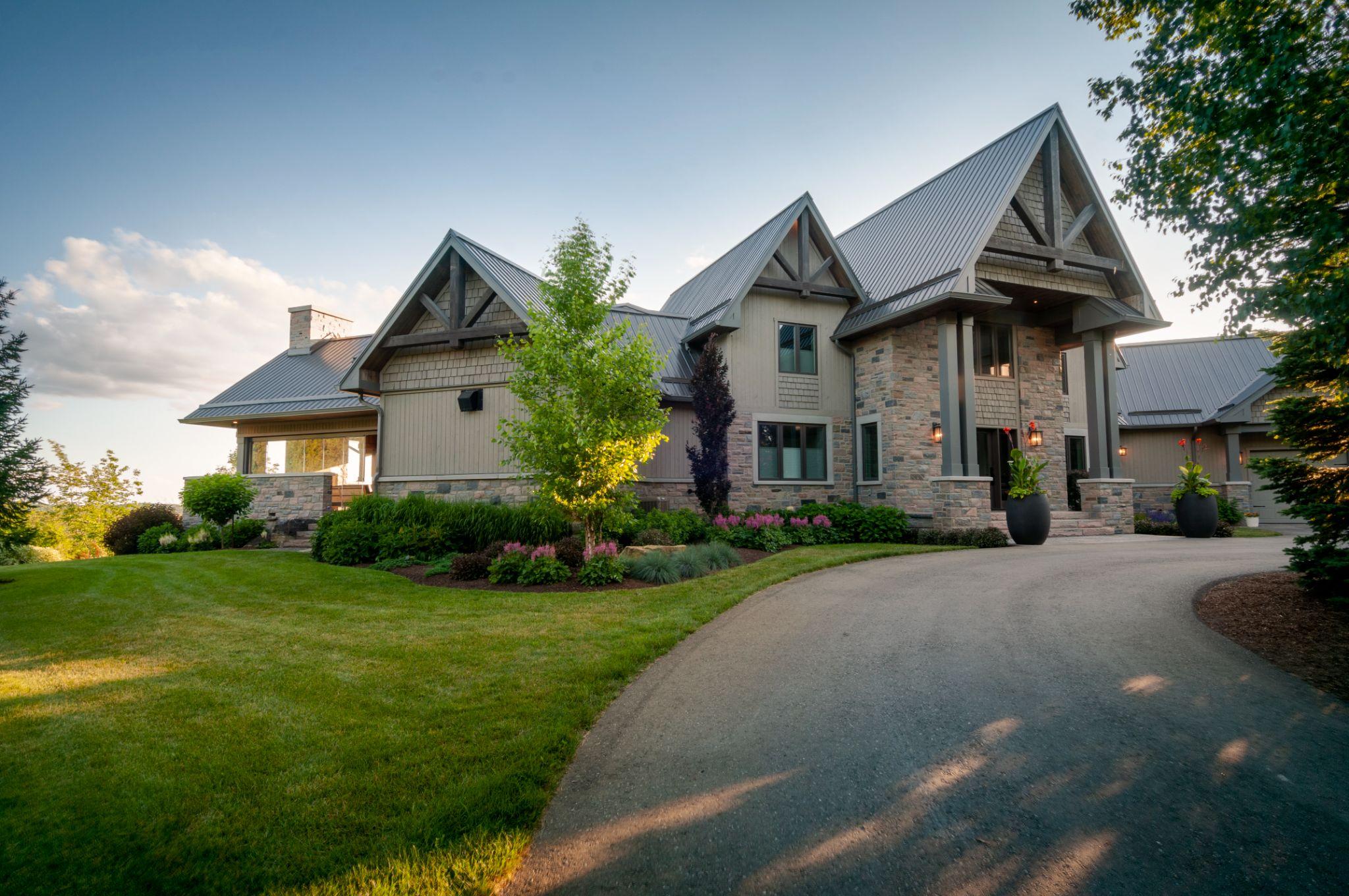 A modern, elegant home with a combination of stone and wood exterior, featuring a steeply pitched roof, large windows, and well-maintained landscaping, set against a clear blue sky.