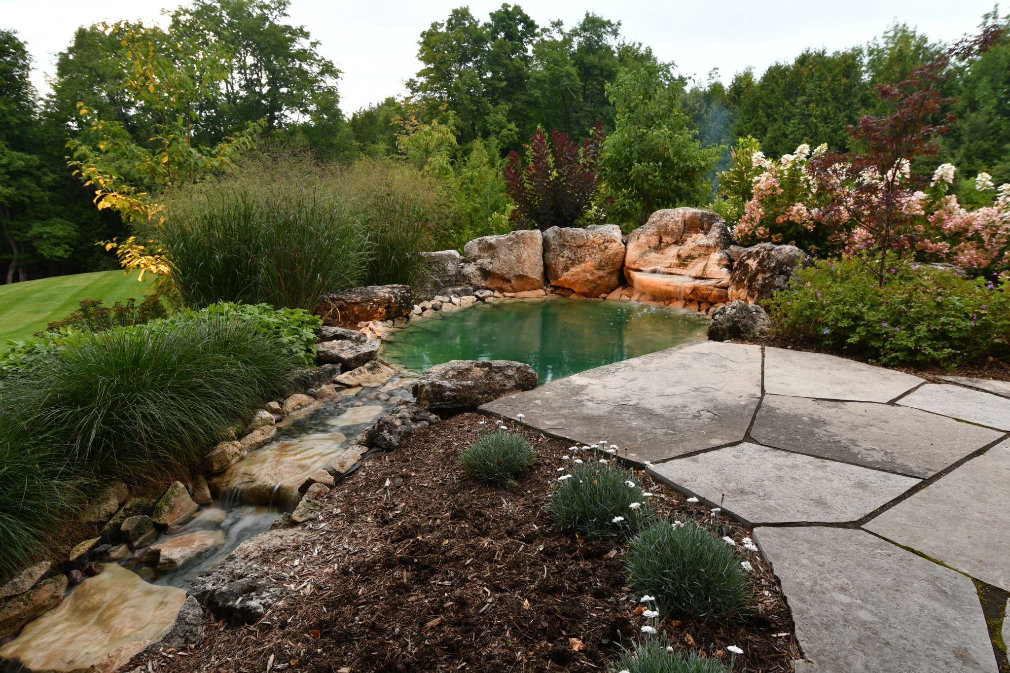 A peaceful garden scene featuring a clear pond surrounded by smooth rocks, a gentle stream flowing nearby, and lush greenery with colorful flowers in the background.