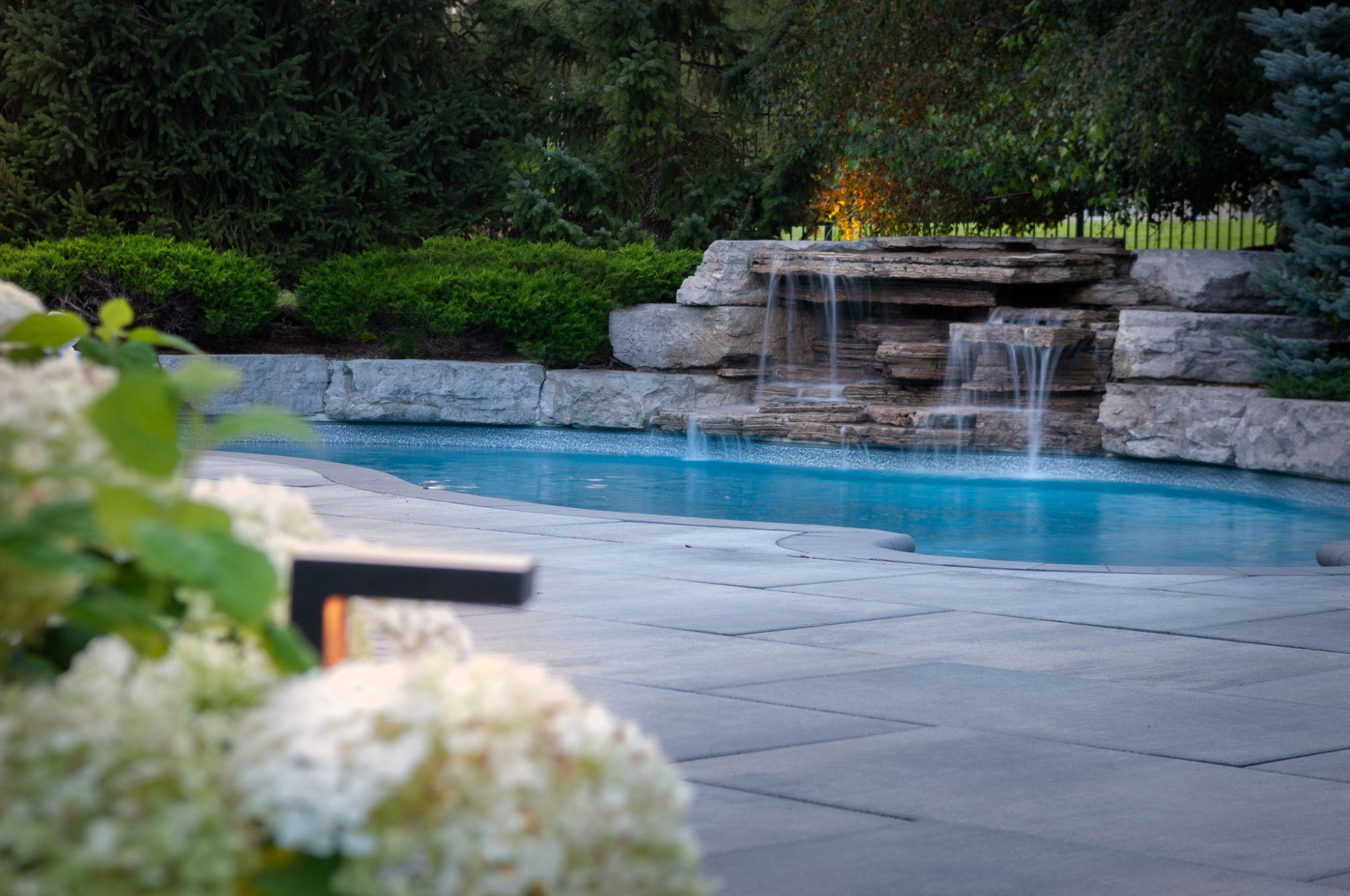 A serene backyard scene featuring a clear blue pool with a natural stone waterfall, surrounded by lush green landscaping and blooming flowers in the foreground.
