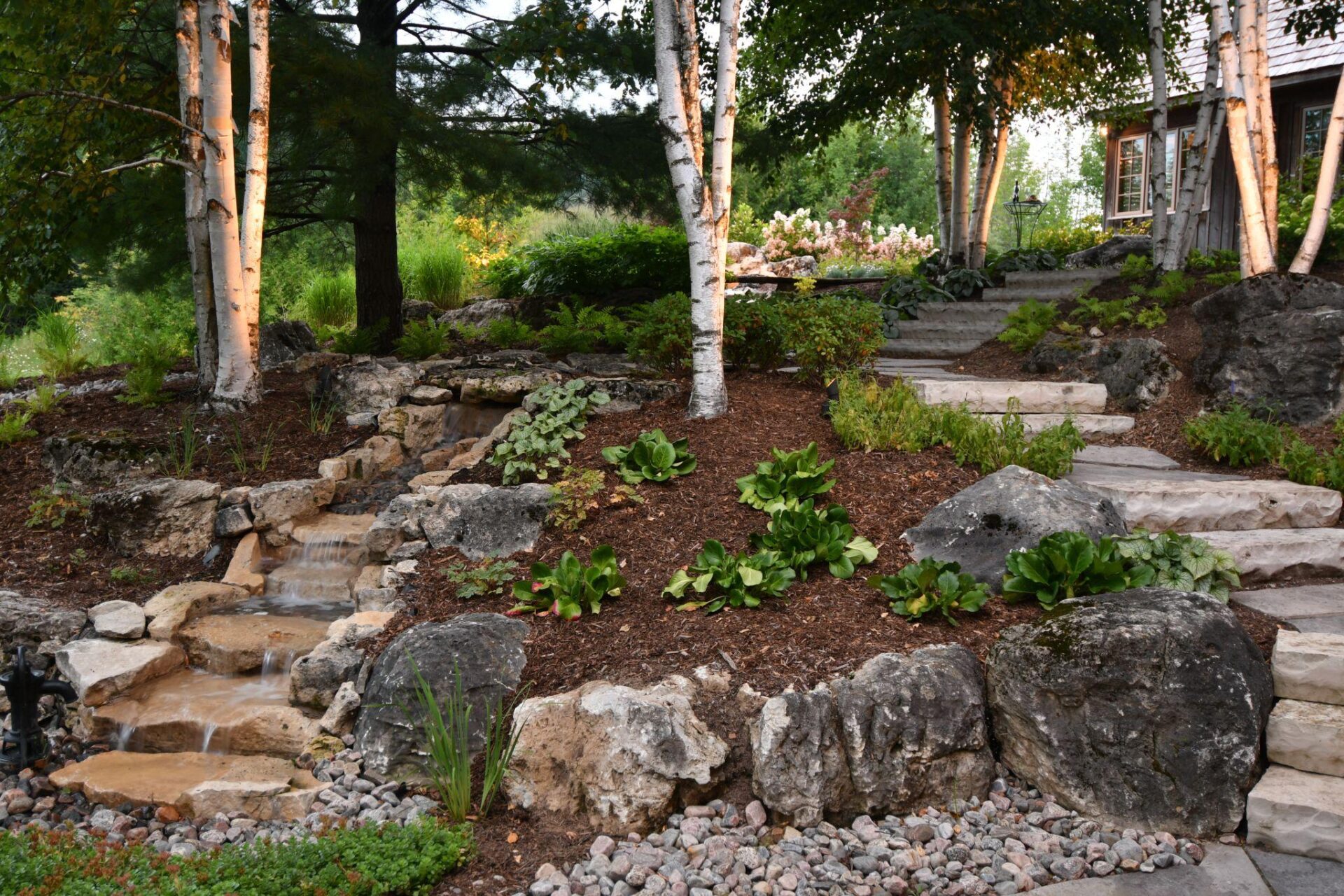 A tranquil garden scene featuring a flowing stream, large rocks, and lush greenery, with birch trees framing the view and colorful plants in the background.