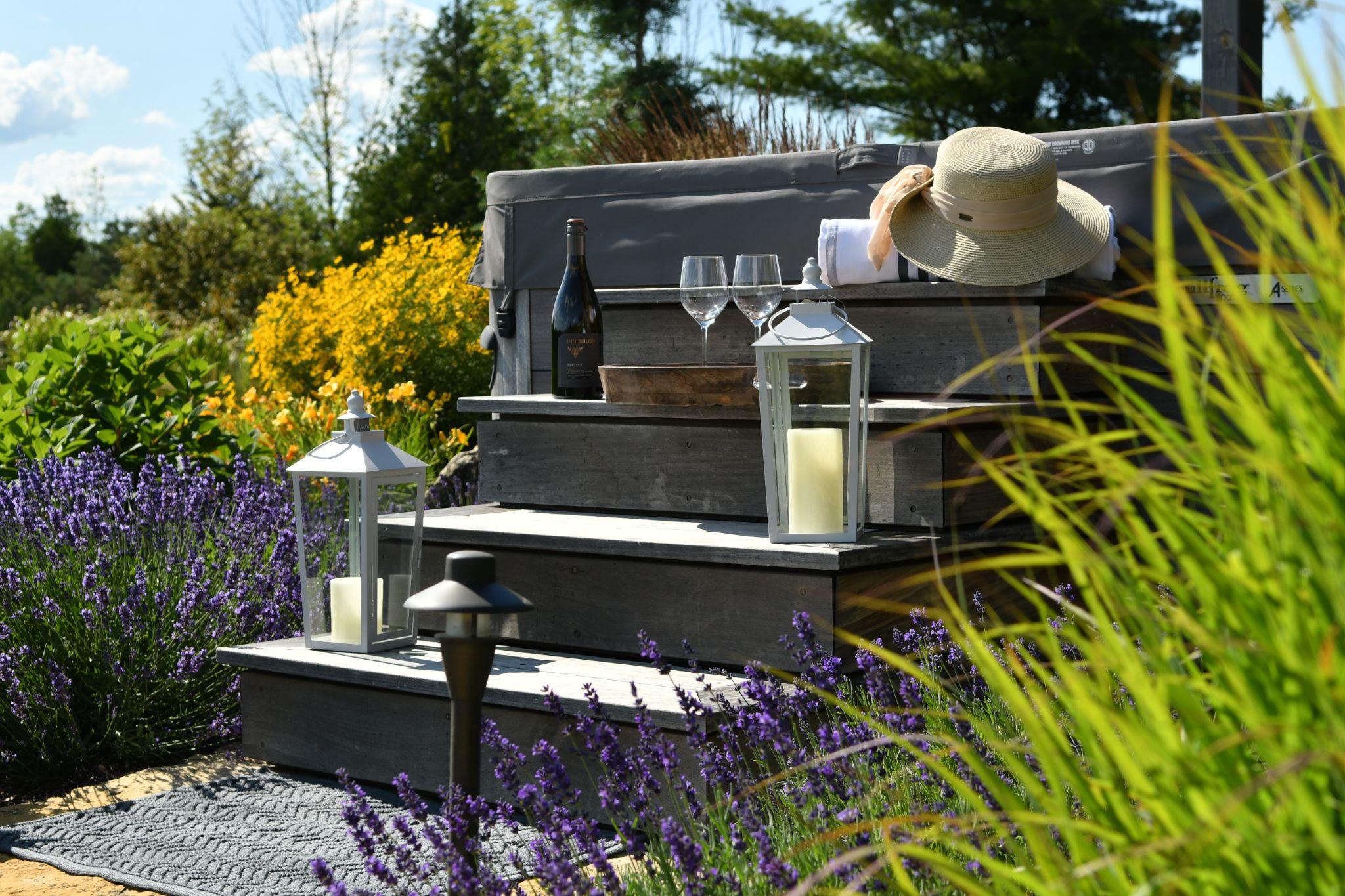 A relaxing outdoor scene featuring a hot tub surrounded by vibrant flowers and greenery. Nearby, there are two glasses of wine on a wooden step, accompanied by a bottle and a decorative lantern, creating a tranquil and inviting atmosphere.
