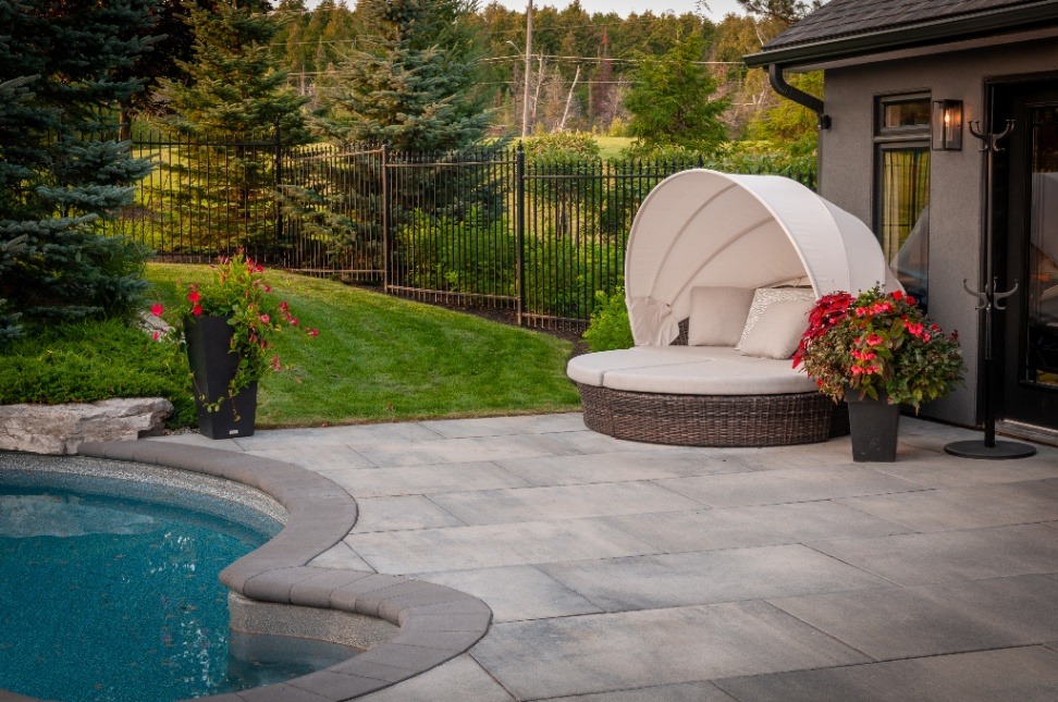 A stylish outdoor lounging area featuring a round, cushioned daybed with a white canopy, surrounded by vibrant flower pots and a sparkling pool, set against a backdrop of lush greenery and a black fence.