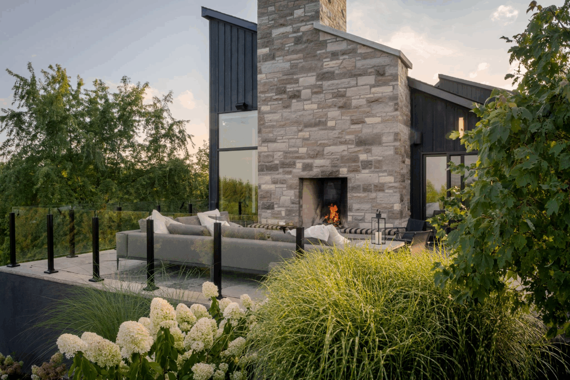 A stylish outdoor lounge area featuring a stone fireplace, surrounded by lush greenery and modern architecture. The comfortable sofa is adorned with cushions, and the warm glow of the fire adds a cozy ambiance, all set against a backdrop of a beautiful sunset.