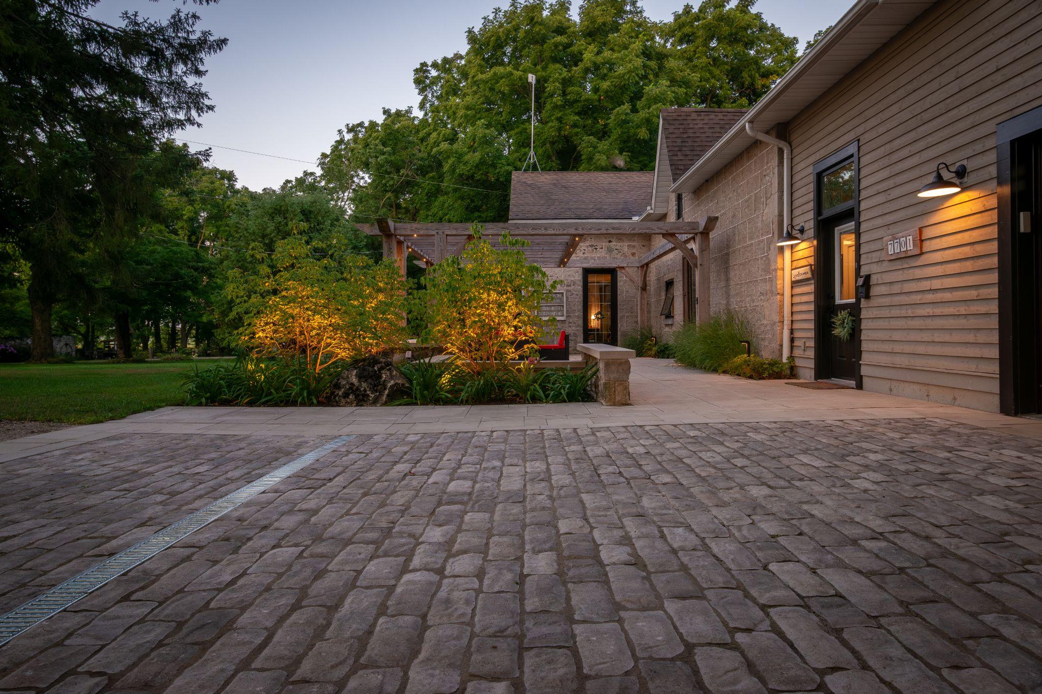Interlock driveway leading and a paver stone path leading to a home with a pergola and warm landscape lighting at dusk.
