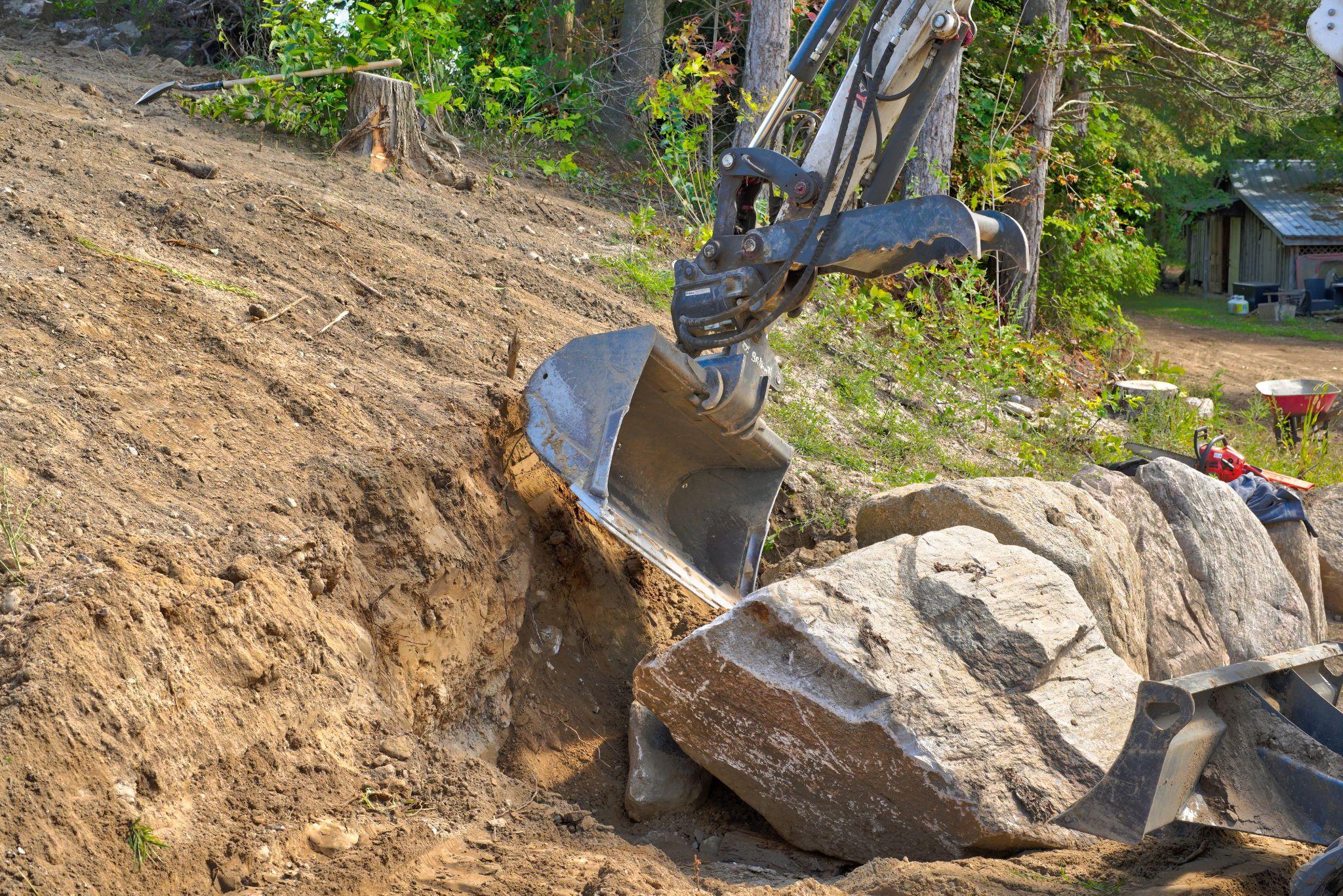 Excavator digging out a large boulder on a wooded hillside at a rural residential property