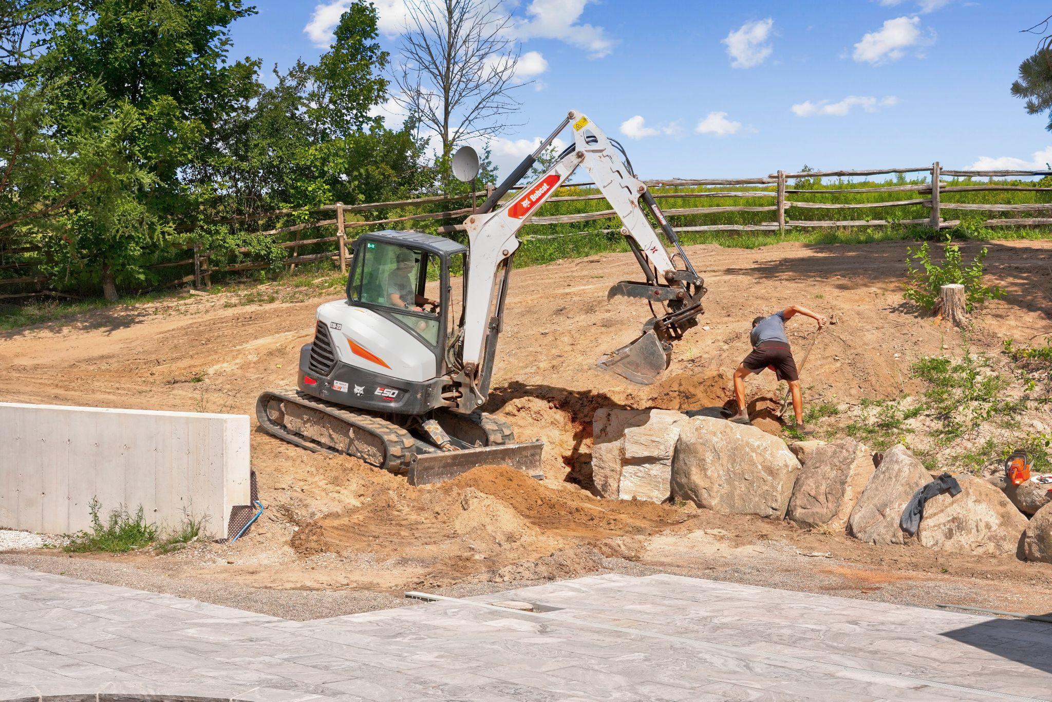 Mini excavator placing boulders during a backyard hardscape installation.