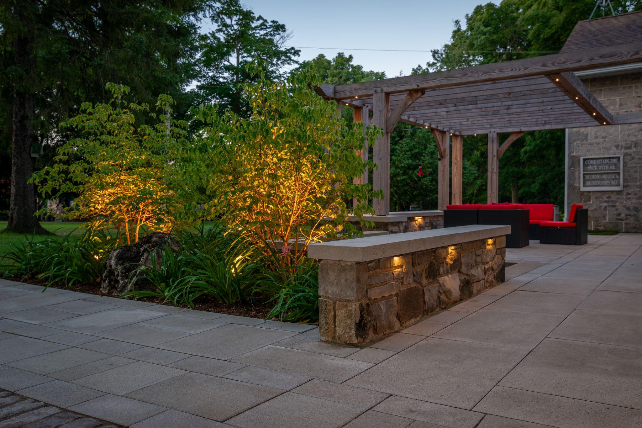 Stone retaining wall with uplit shrubs beside a paver patio and red-cushioned outdoor seating at dusk.